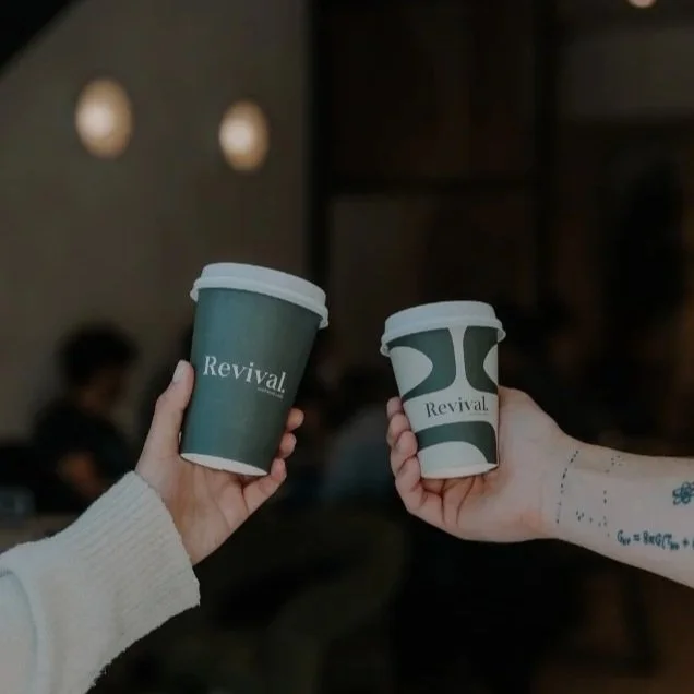Two people holding takeaway coffee cups with the word "Revival" on them, in a dimly lit indoor setting.