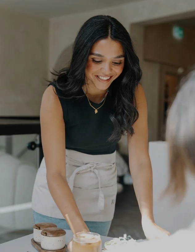 A woman smiling and leaning over a table with desserts, wearing a black sleeveless top and a beige apron, in a cozy indoor setting.