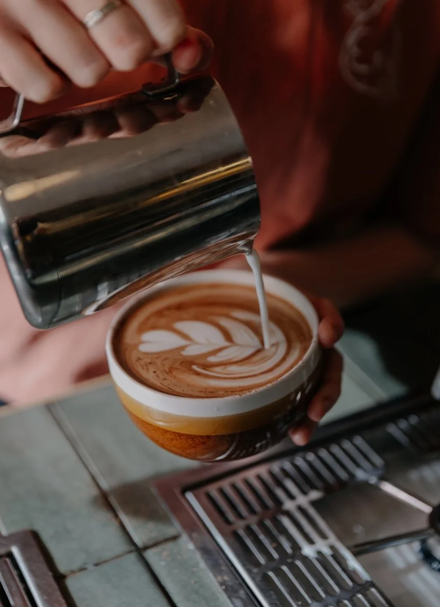 A barista pours steamed milk into a cup of espresso to create latte art, with the barista holding a metal pitcher and the cup being held with one hand. The latte art appears as a leaf pattern.