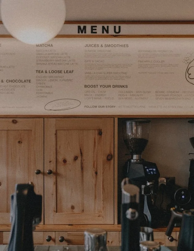 Tapas-style restaurant menu board displaying a list of matcha, juices & smoothies, tea & loose leaf, and chocolate drinks, mounted above a wooden cabinet in a cozy cafe setting.