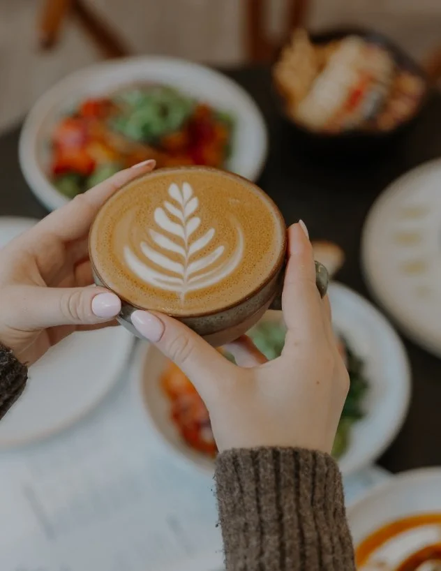 A person holding a cup of latte with latte art, surrounded by various dishes on a table.