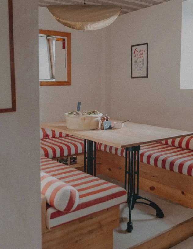 A cozy dining nook with a wooden table surrounded by built-in benches with red and white striped cushions. There is a bowl of food on the table, a small sign, and a mirror on the wall. The space has light-colored walls and a small framed poster, with a round hanging light fixture above.