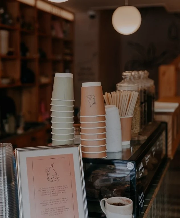 Stacked disposable coffee cups on a counter at a coffee shop, with a framed sign and a cup of coffee, against a background of shelves and warm lighting.