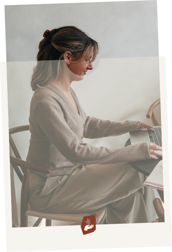 A woman sitting at a desk using a laptop with a notebook nearby.