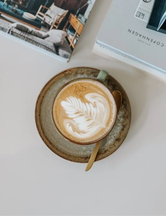 A cup of coffee with latte art on top, placed on a saucer with a spoon, on a white table next to a magazine and a catalog.