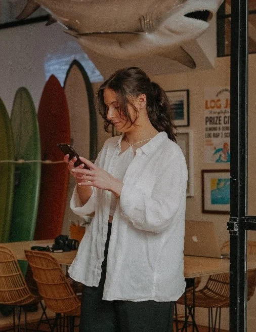 Young woman with long wavy hair, wearing a white jacket, using her cellphone inside a beach-themed restaurant with surfboards on the wall and framed pictures.