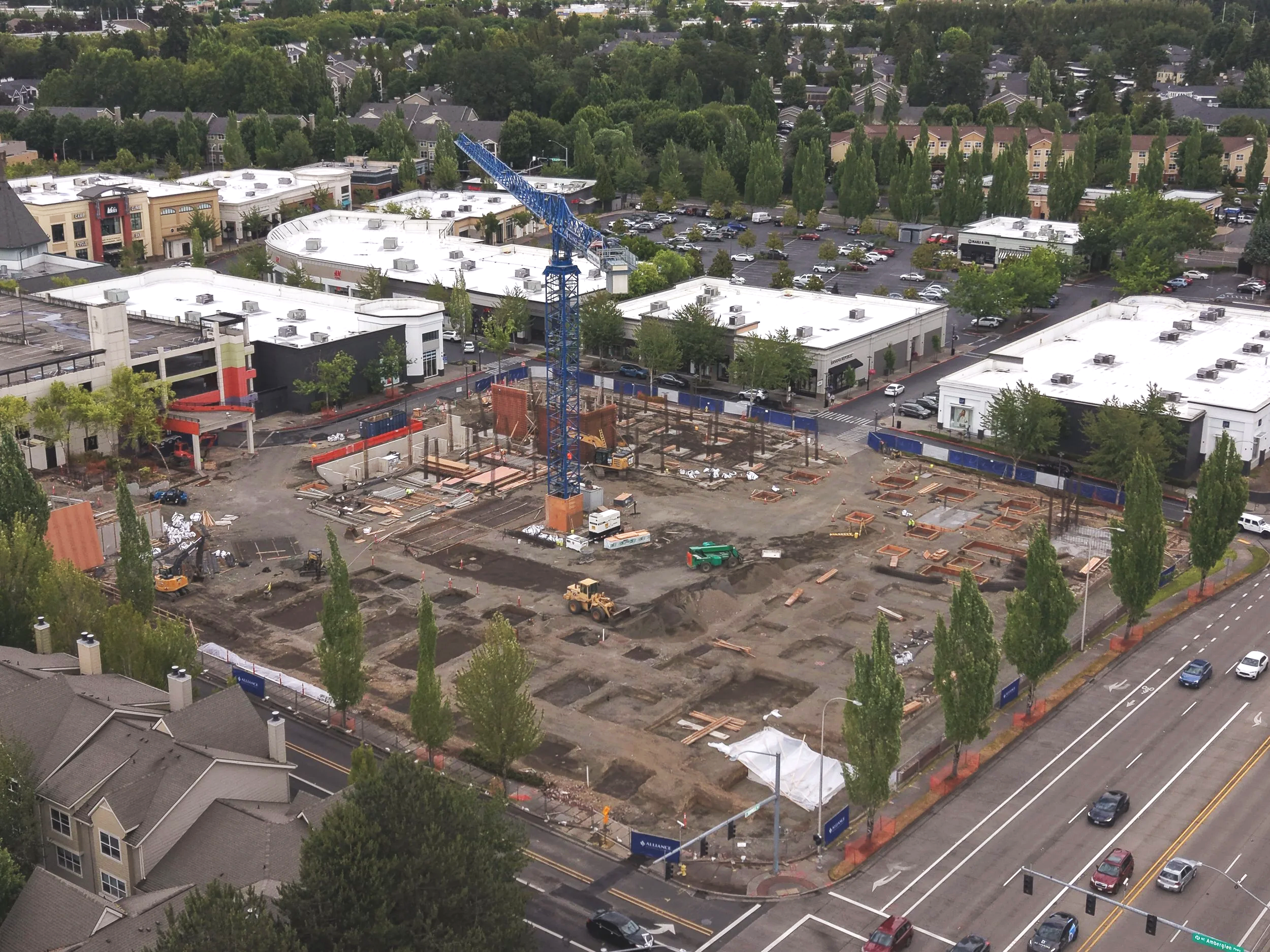 An aerial view of a shopping center under construction, with a blue crane, construction site, and surrounding buildings and parking lots.