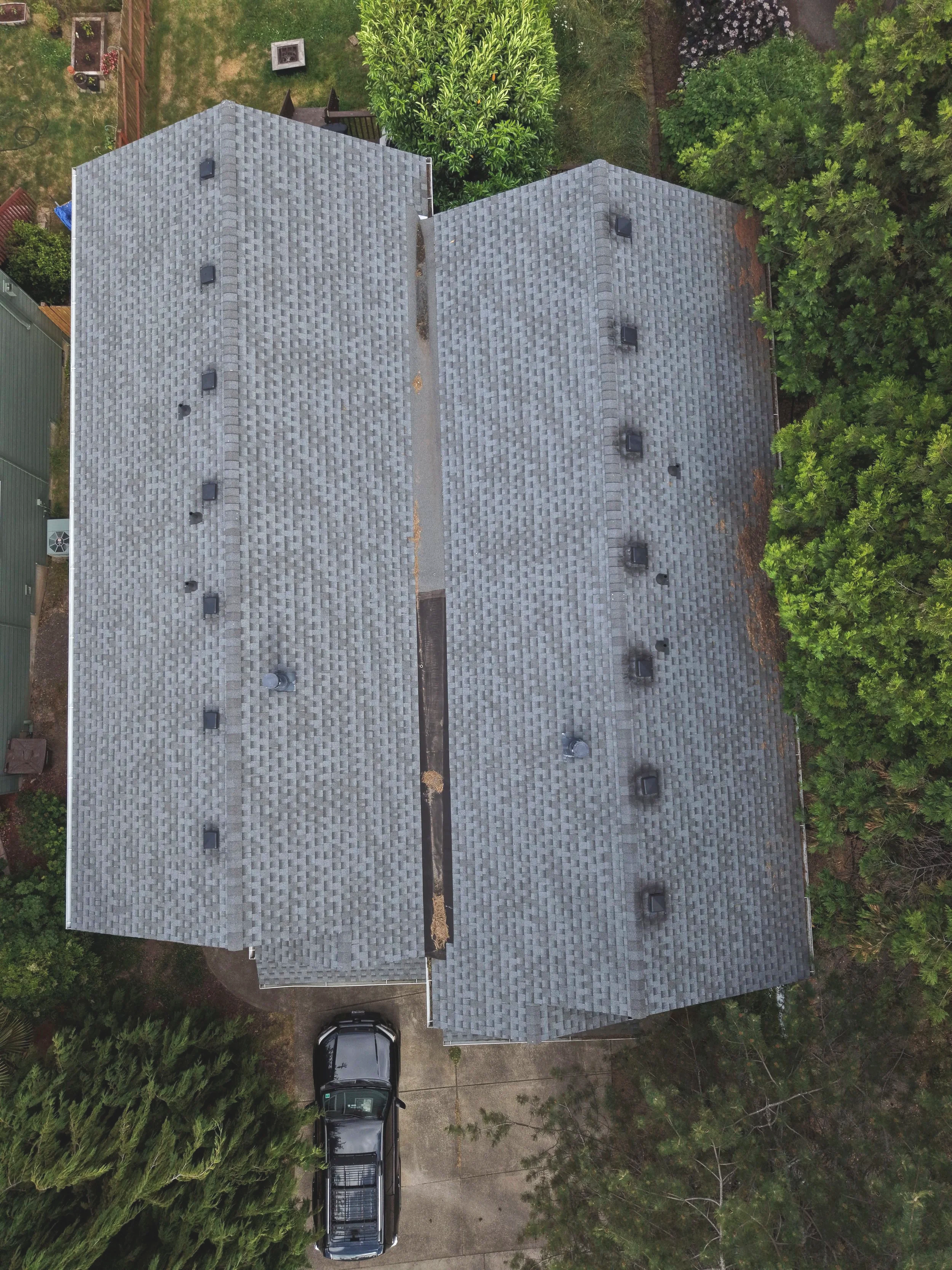 Aerial view of a house with gray shingle roof, surrounded by green trees and a driveway with parked cars.