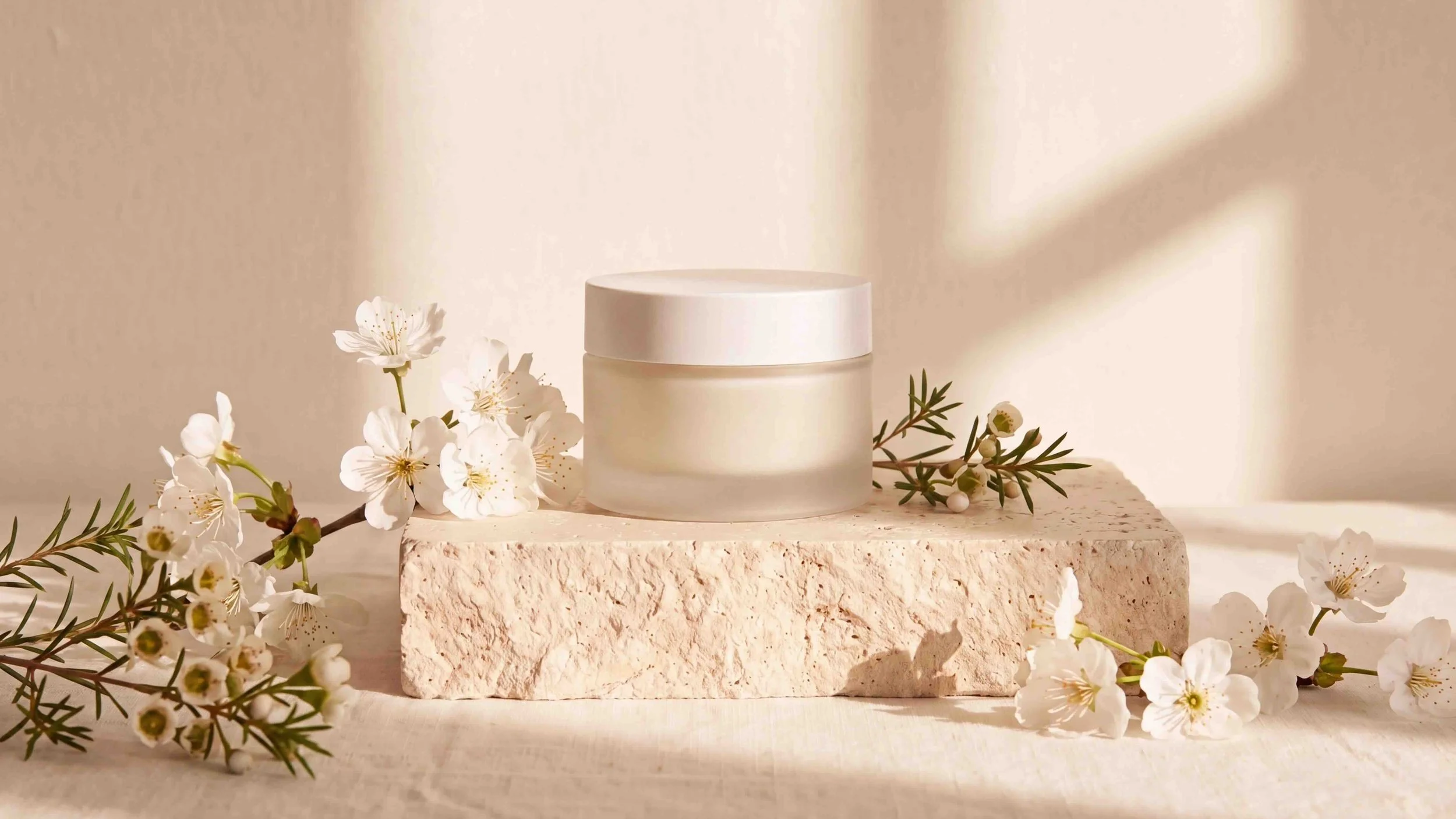 A white jar cosmetic container on a textured stone block, surrounded by white flowers and greenery, with a neutral beige background and soft lighting.