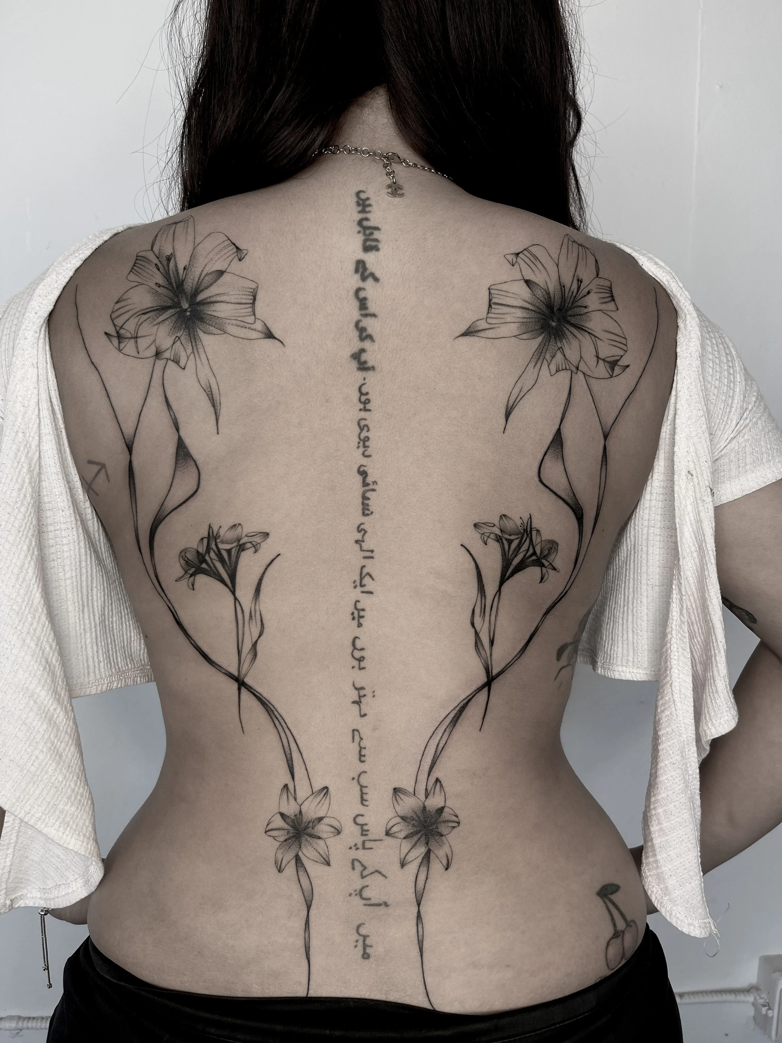 A person's back with a black and gray tattoo of lilies and a vertical line of script text running down the center of the back.