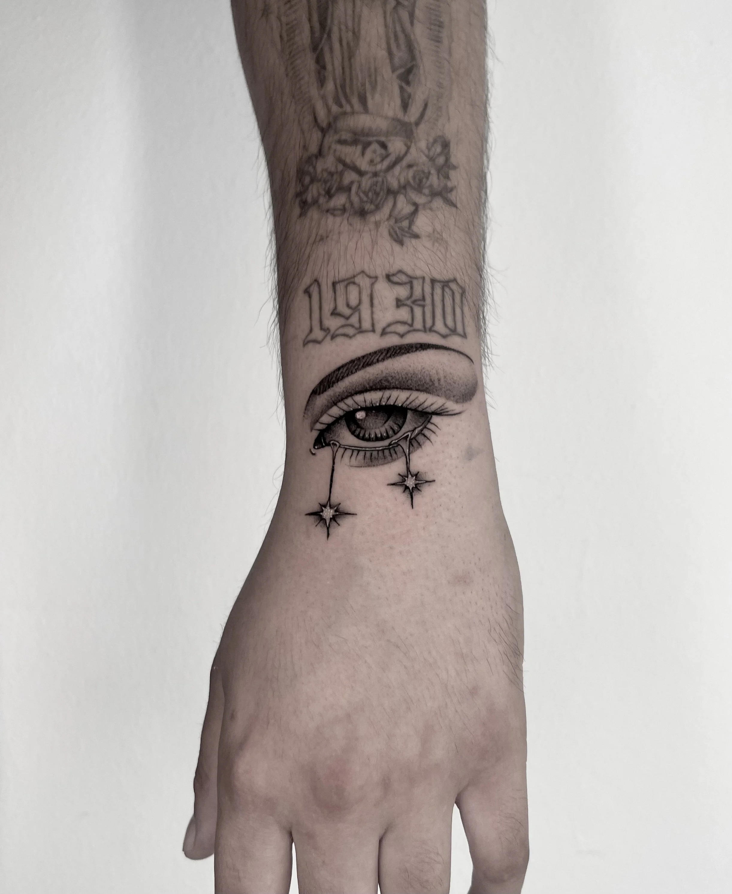 A close-up of a tattooed arm and hand against a white background. The tattoos include an eye with a decorative design, the years 1970-2020, and some flowers and other decorative elements.