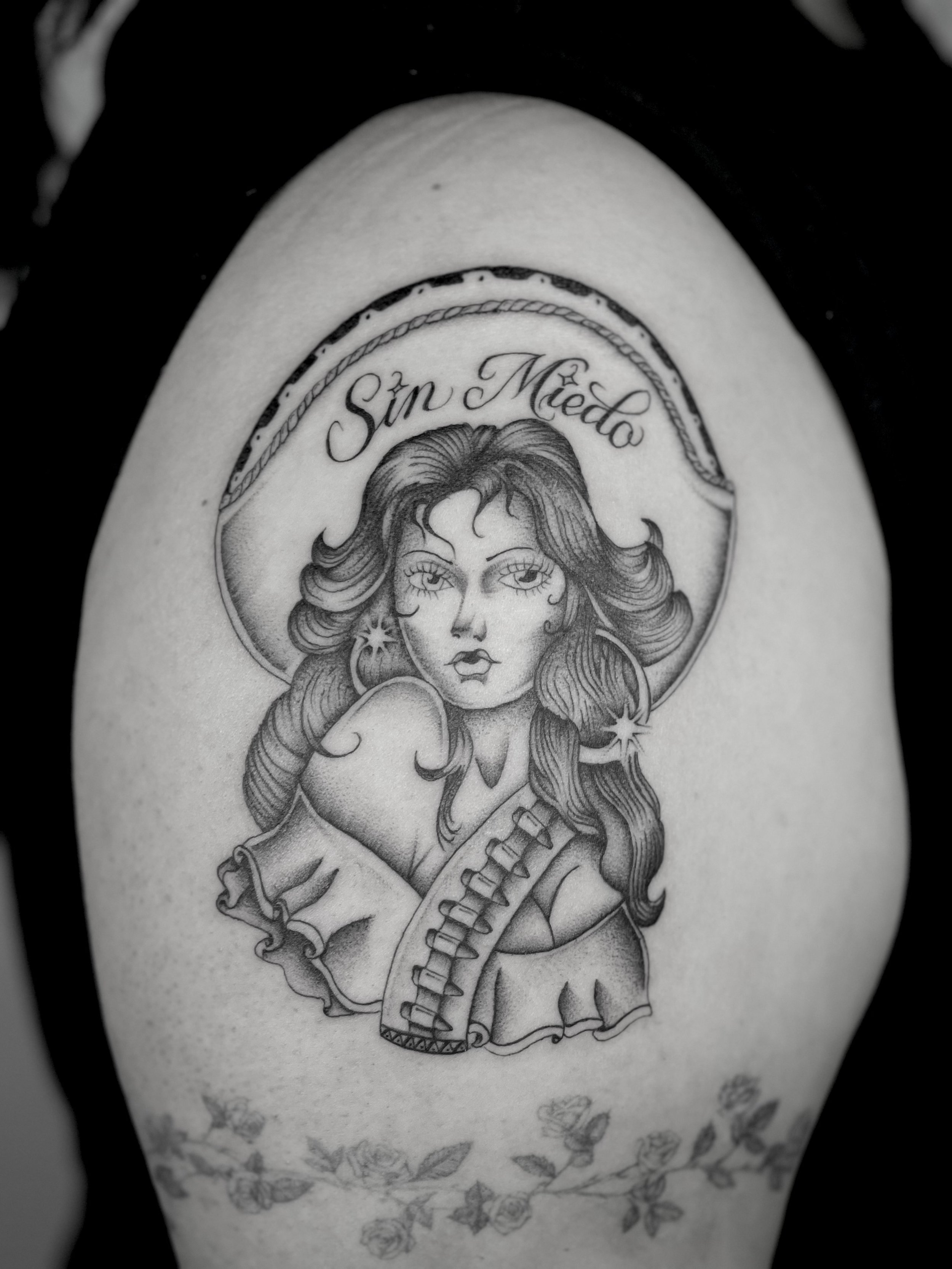 Black and white tattoo of a woman with long, wavy hair and a halo, with text "Sin Miedo" above her head, surrounded by a decorative border.