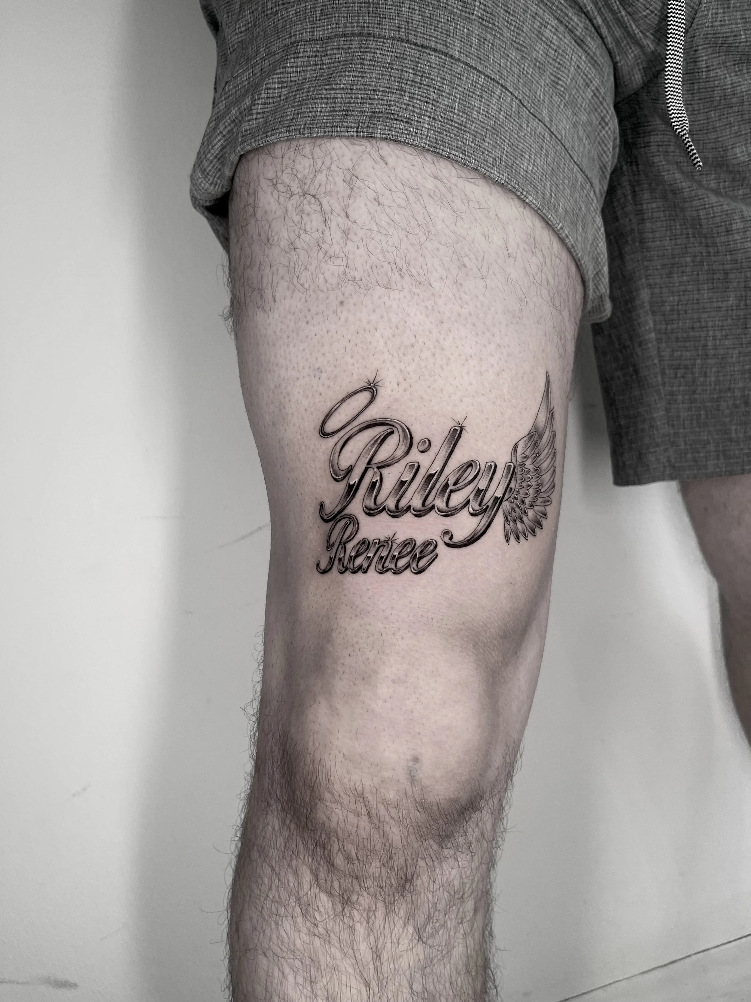 A person's upper leg and part of their shorts, revealing a tattoo on the thigh with the words 'Riley Renee' and a winged design.