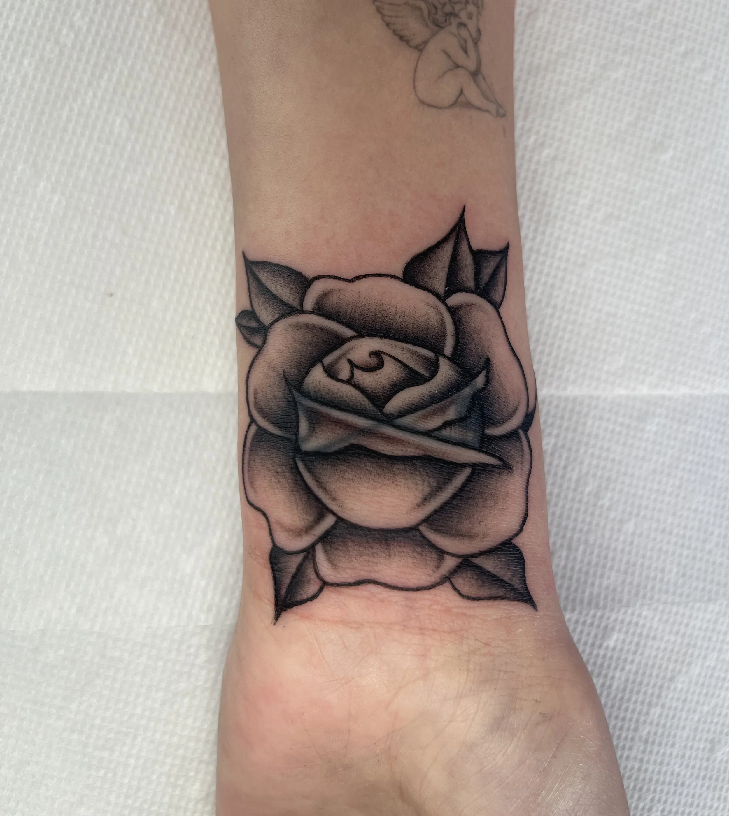 A tattoo of a rose on a person's arm, with a smaller ink drawing of a cherful baby with wings above it.