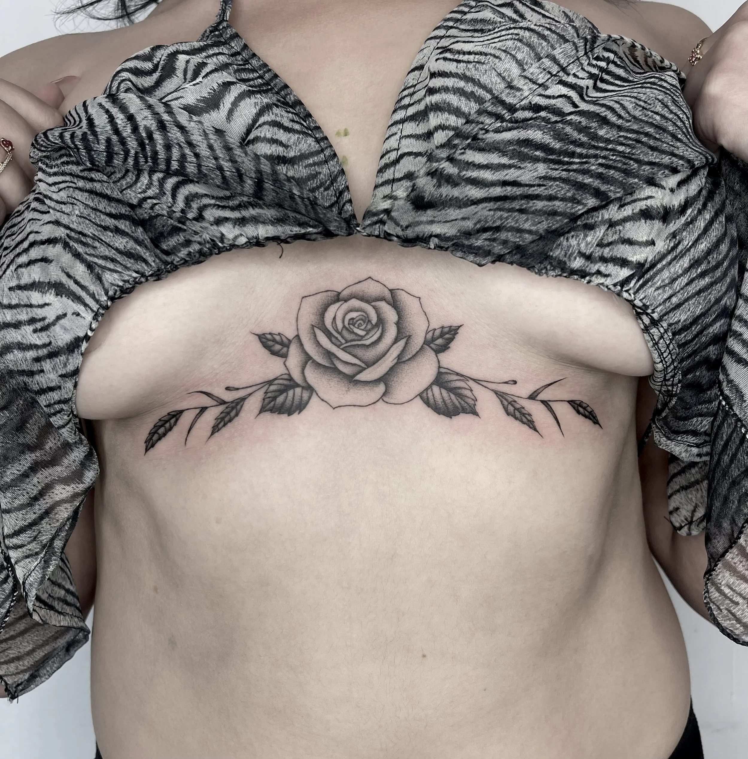 A woman wearing a zebra-print top, showing part of her chest with a tattoo of a rose and leaves, and hands holding the fabric.