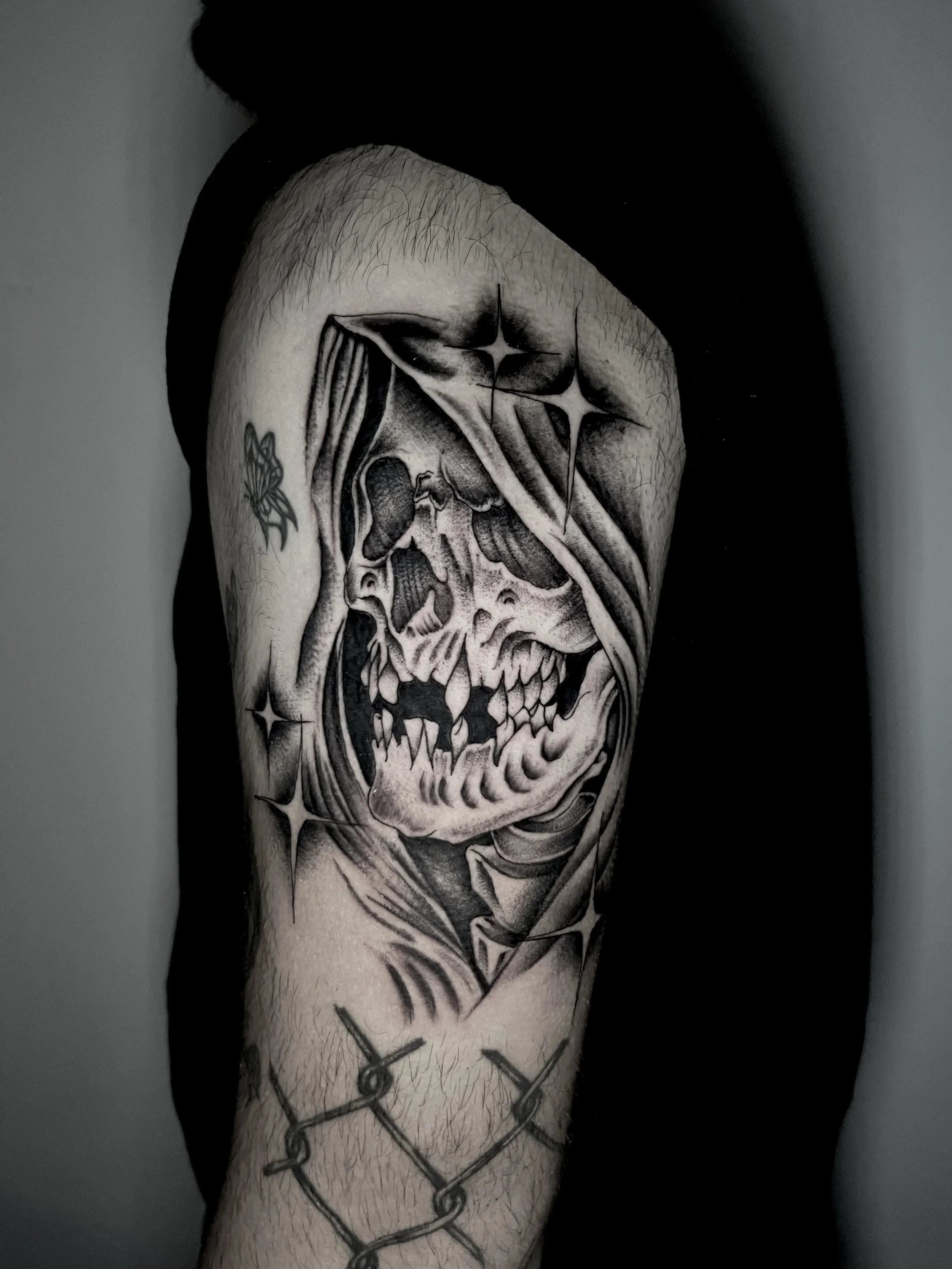 Black and gray tattoo of a stylized skull with some stars and abstract elements on a person's upper arm. The person is wearing a black short-sleeve shirt.