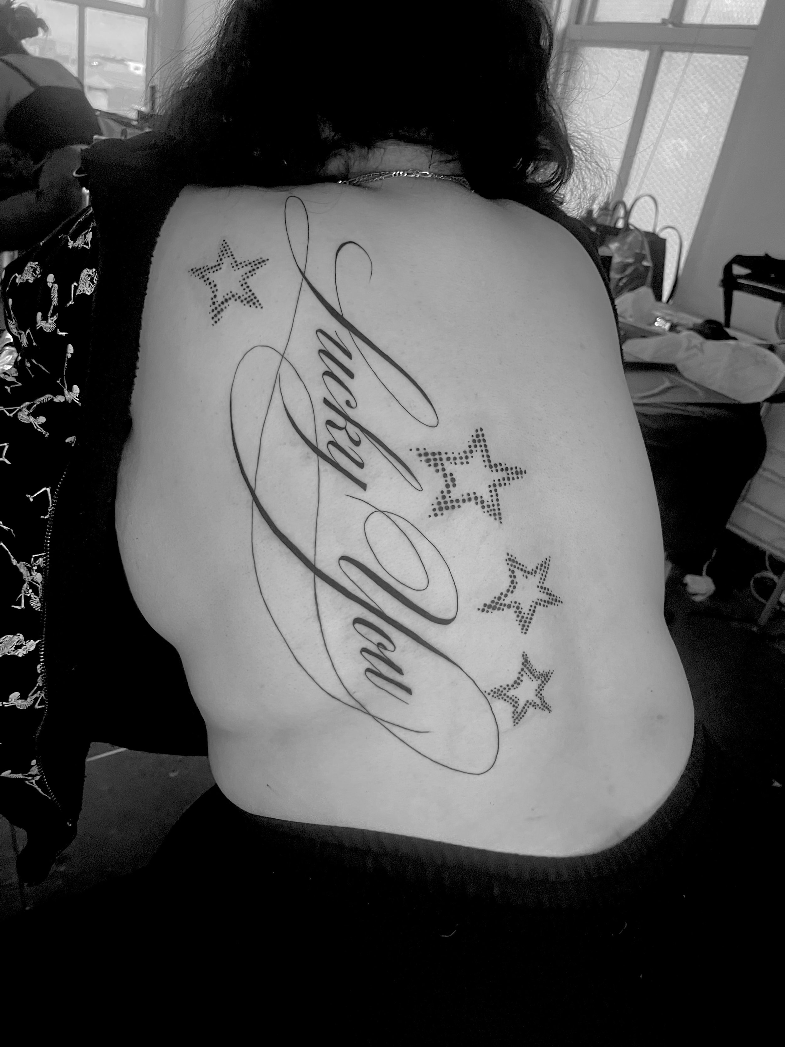 Back of a person's shoulder and upper back with a tattoo that says 'MOM' cursively, surrounded by five star designs composed of small dots, with black curly hair and a dark shirt. The background shows a room with a window, some bags, and objects.