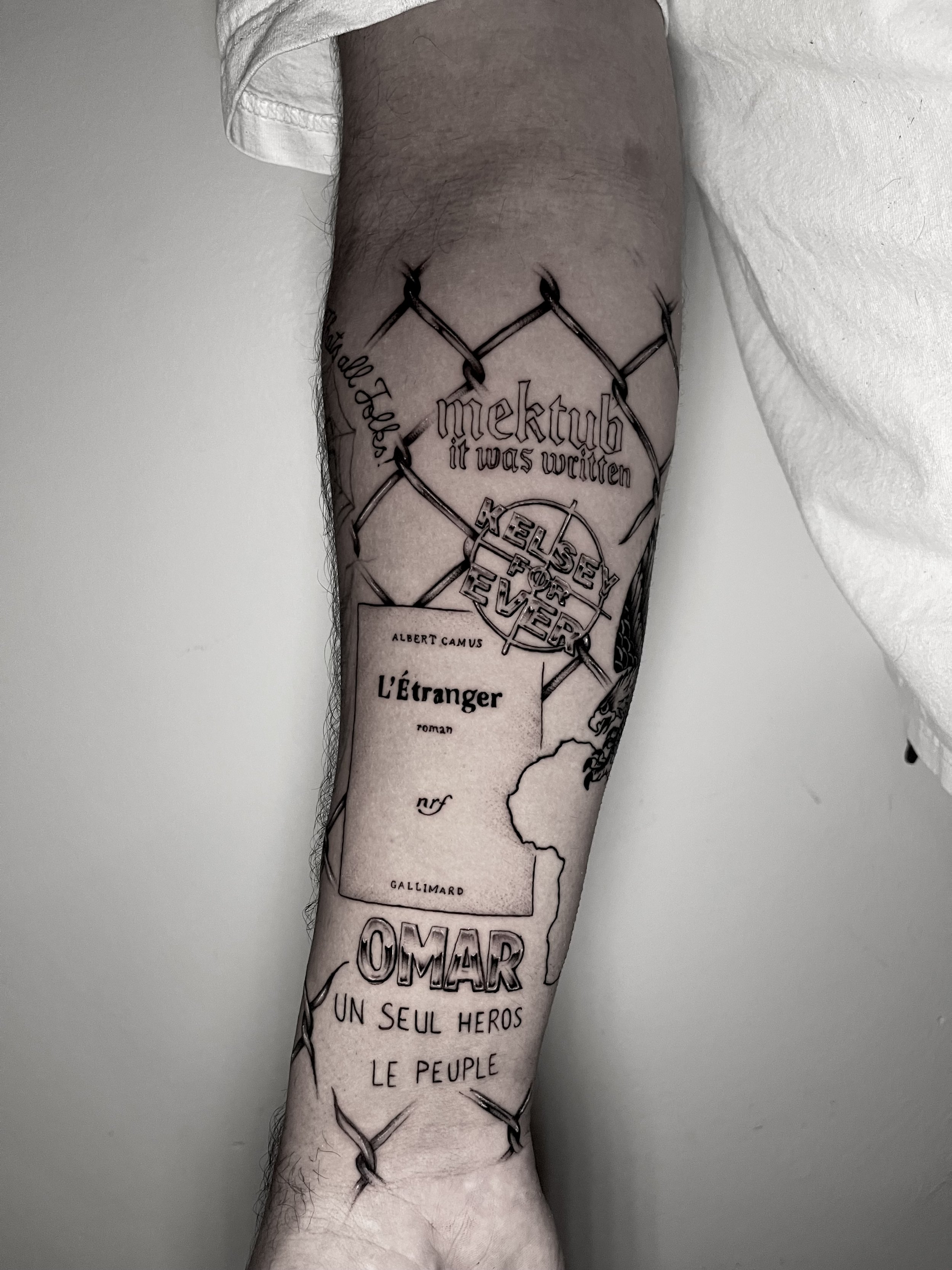 Tattoo on a person's arm featuring a chain-link fence with the text "mektub it was written" and a map outline of South America with various place names and French words.