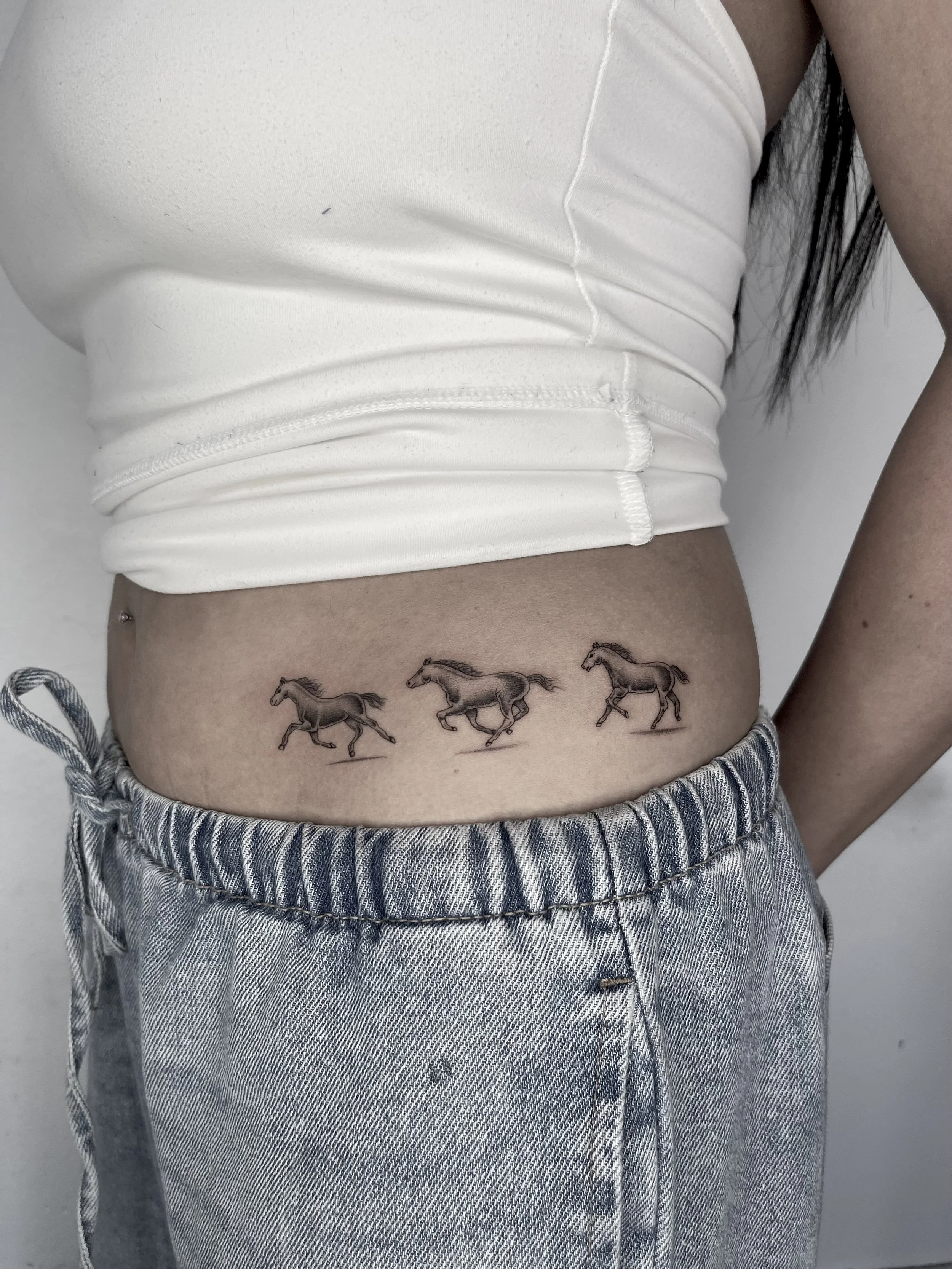 Close-up of a person's torso wearing a white sleeveless top and blue and white striped pants, with a tattoo of three running horses on their lower abdomen.