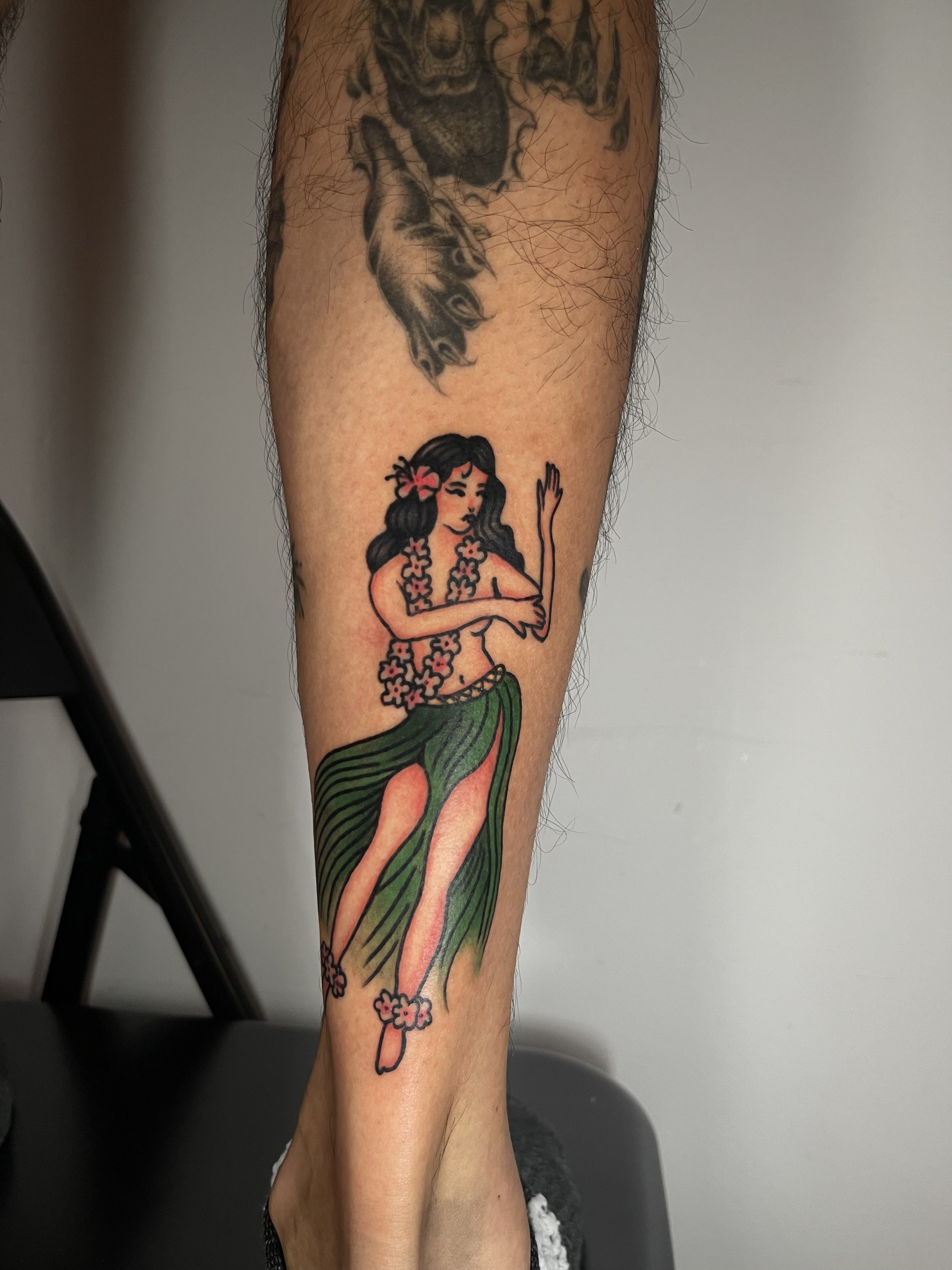 Tattoo of a hula girl with a pink flower in her hair, wearing a floral lei and a green hula skirt, on a person's arm.