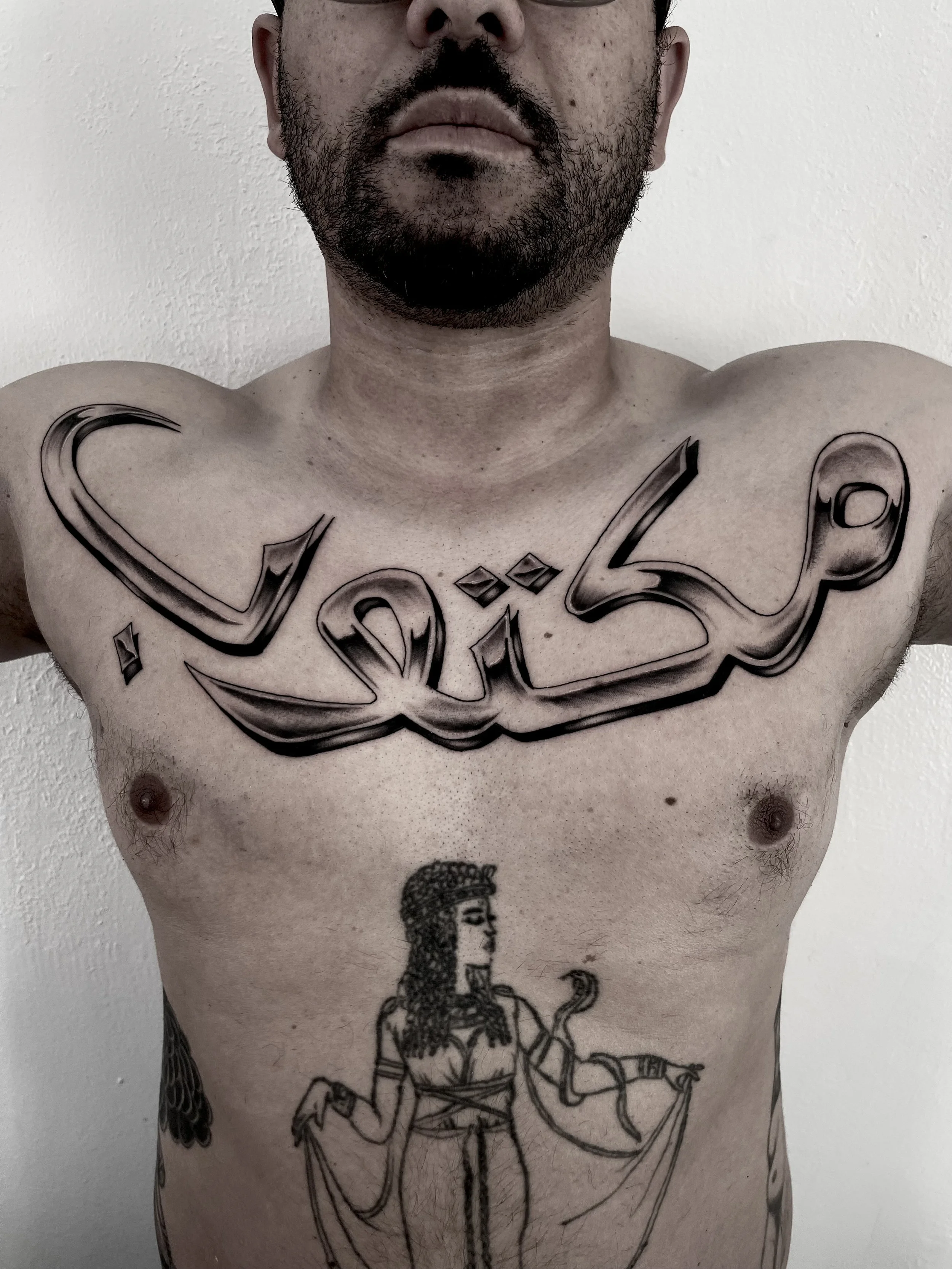 A shirtless man with tattoos on his chest and torso, including a large stylized text tattoo across his chest and an illustration of a woman below. The man has a beard and mustache, and his face is partially visible in the image.