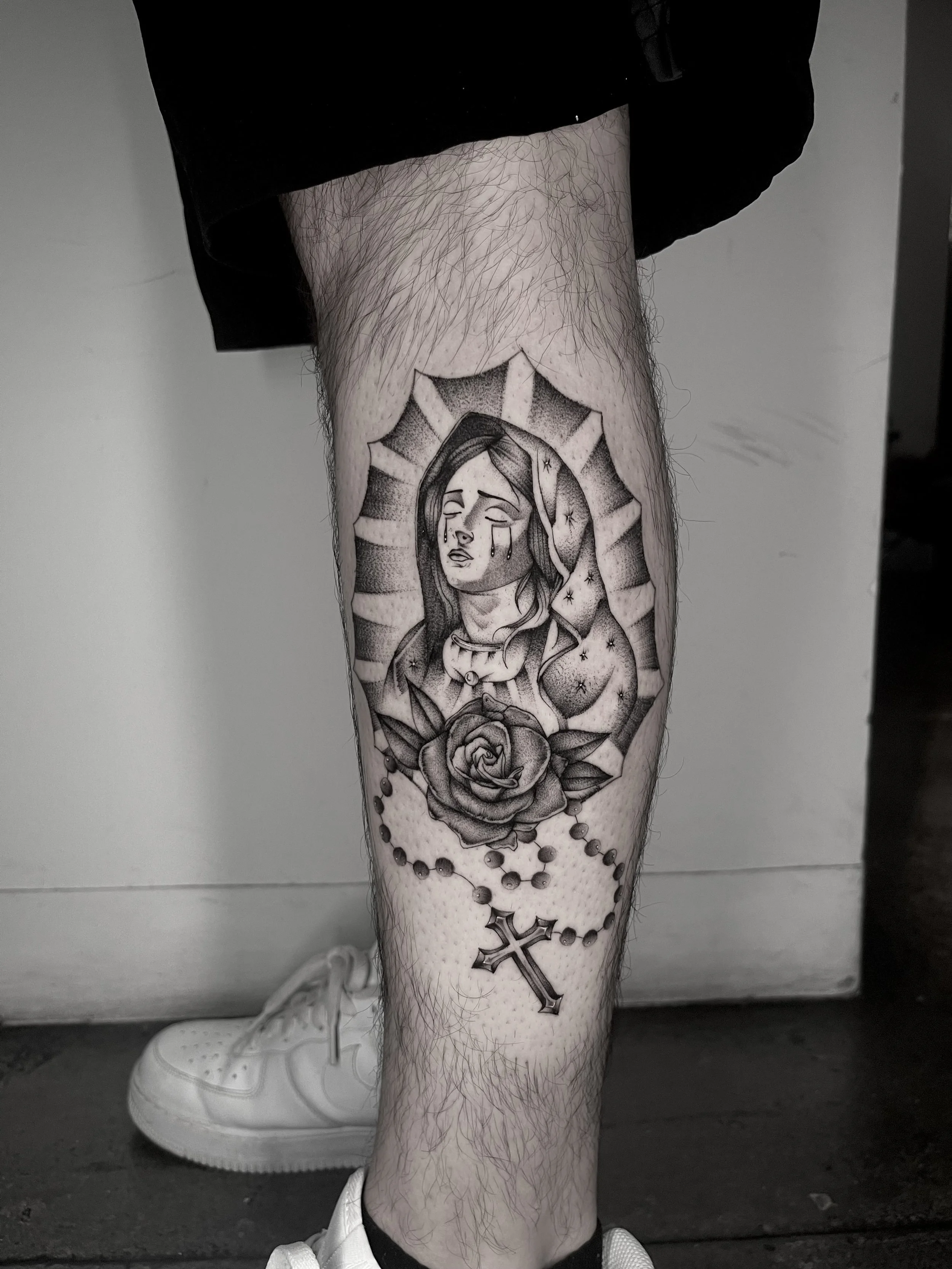 A person's leg with a tattoo of a crying woman, a rose, and a cross.