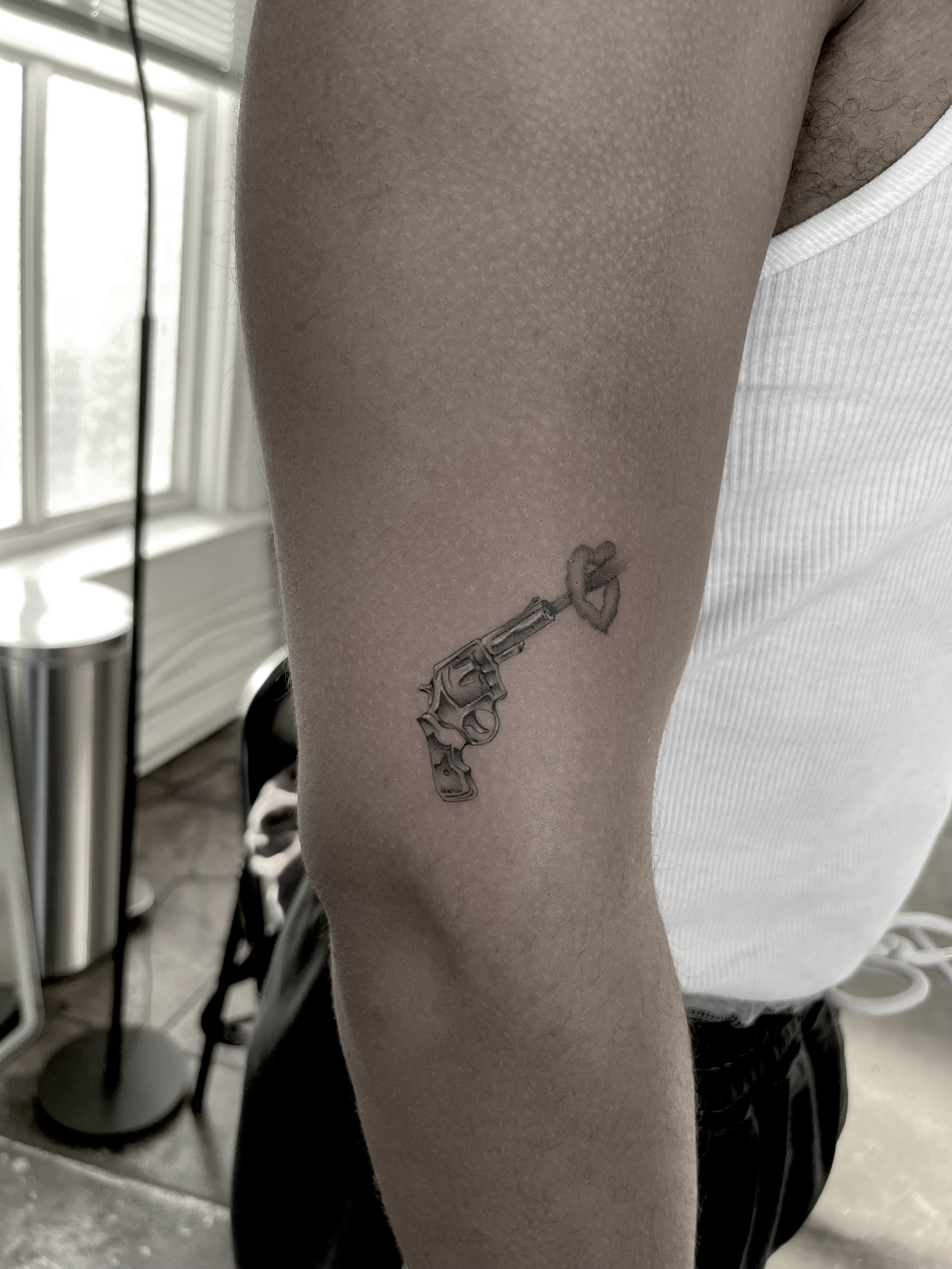 Close-up of a person's thigh with a tattoo of a gun and a heart. The person is wearing a white sleeveless shirt and black pants, standing indoors with a window and furniture in the background.