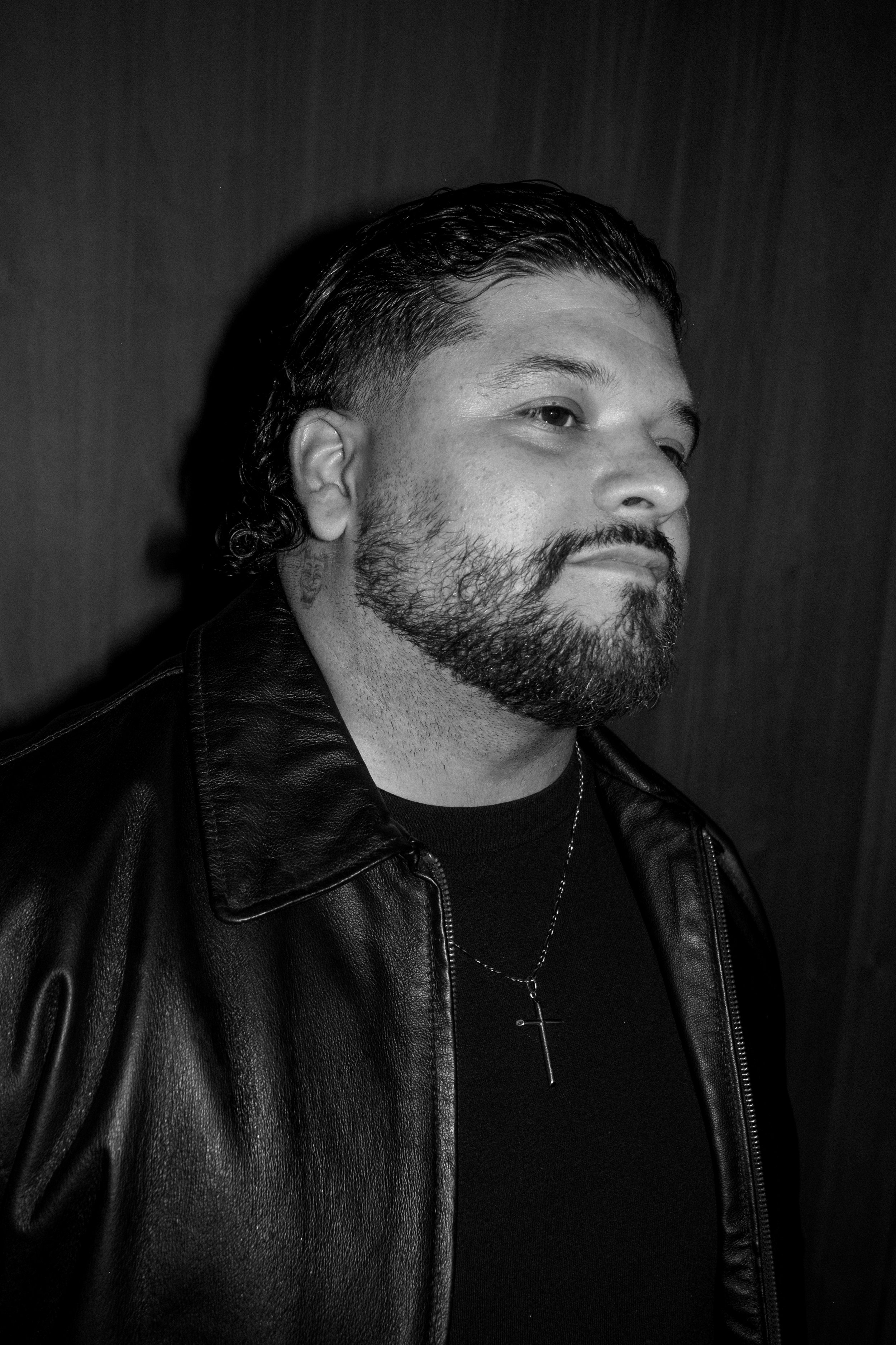 Black and white photo of a man with a beard and dark hair, wearing a leather jacket with a zipper and a chain necklace, looking to the right against a dark background.