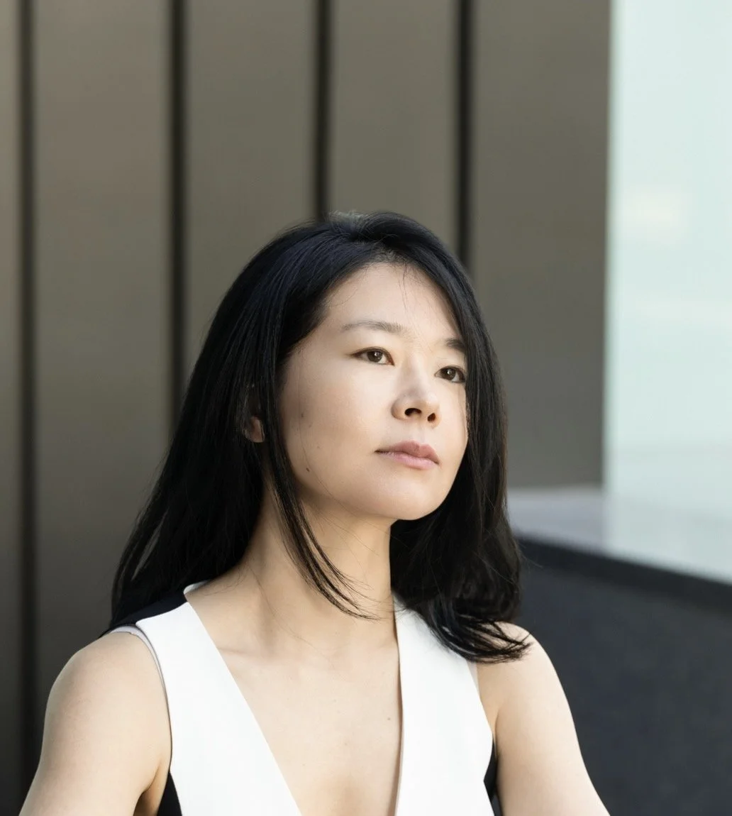 A woman with straight black hair, wearing a white sleeveless top, standing outdoors against a dark wooden and light-colored background.