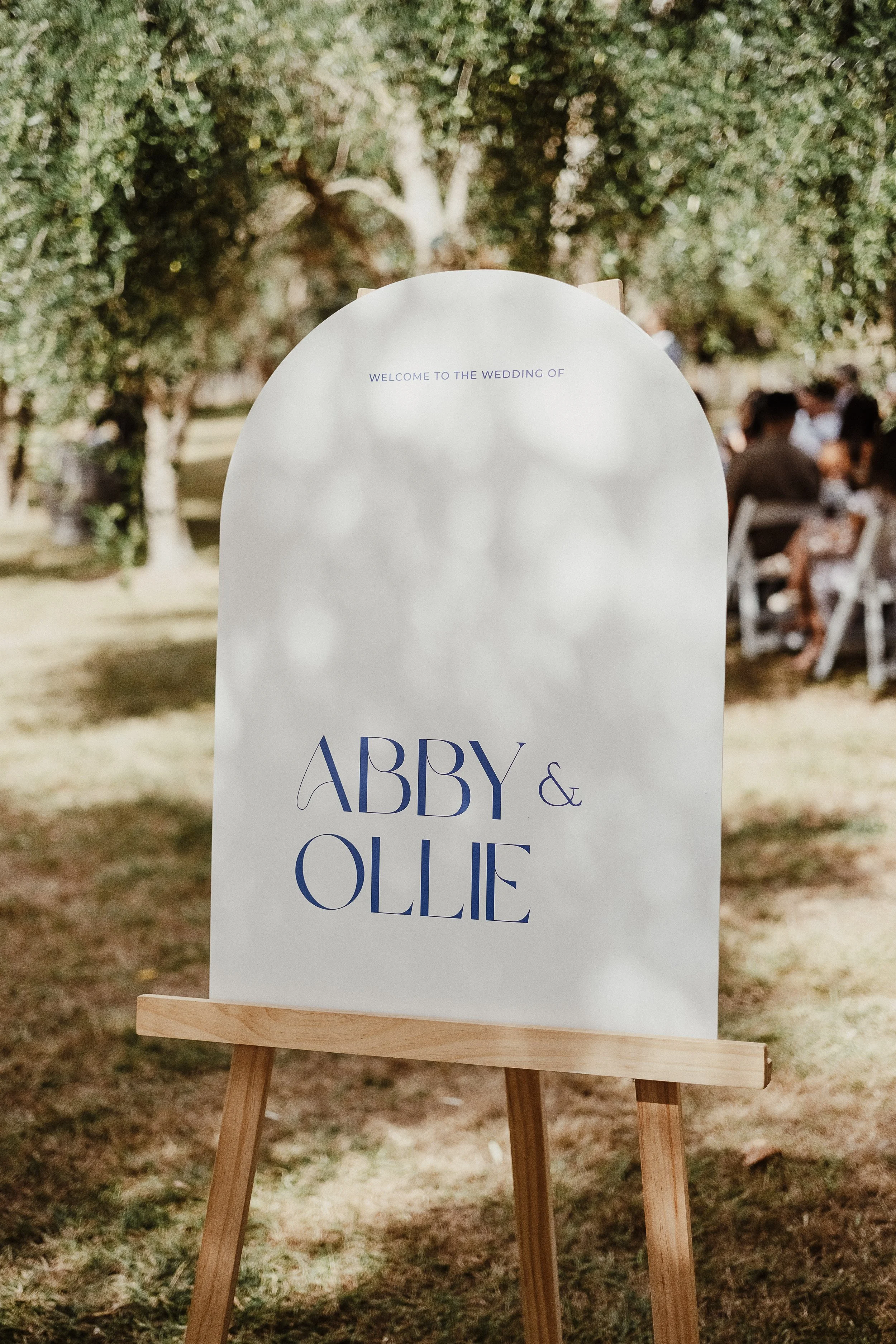 Wedding sign with the names Abby and Ollie, standing on a wooden easel outdoors with trees and guests in the background.