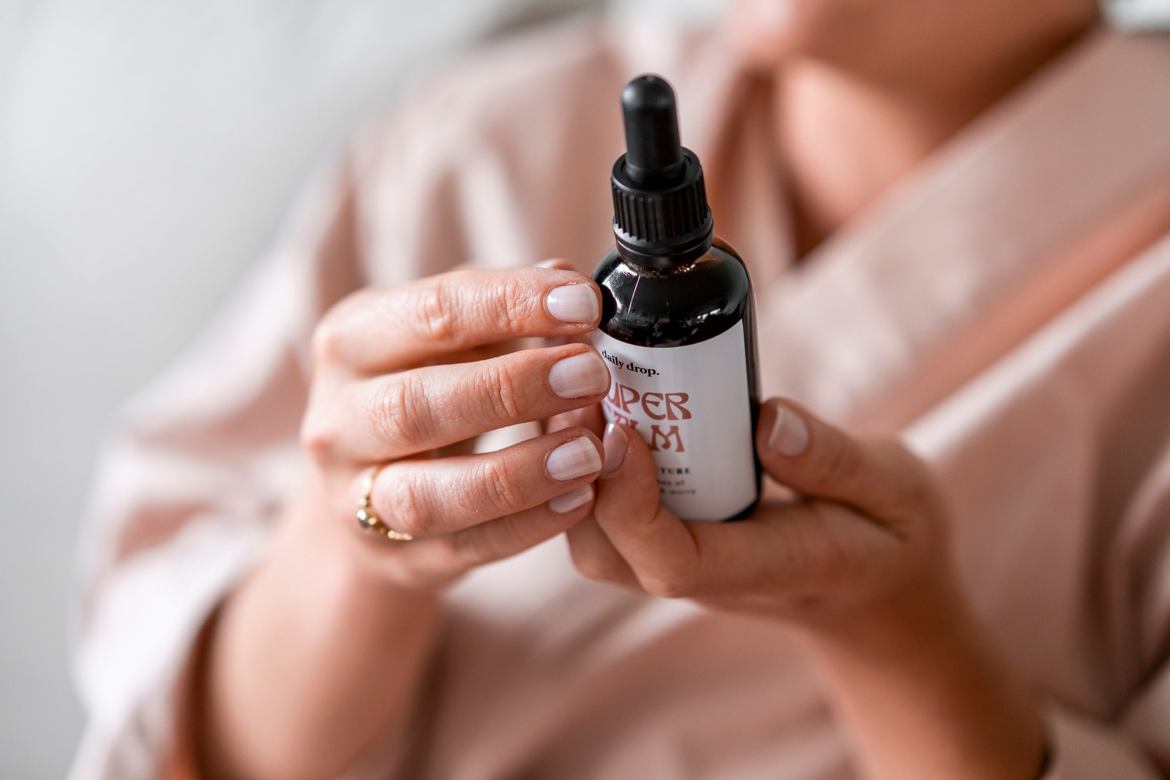 Close-up of a person holding a small dark bottle with a black dropper cap, with their other hand slightly blurred in the background.