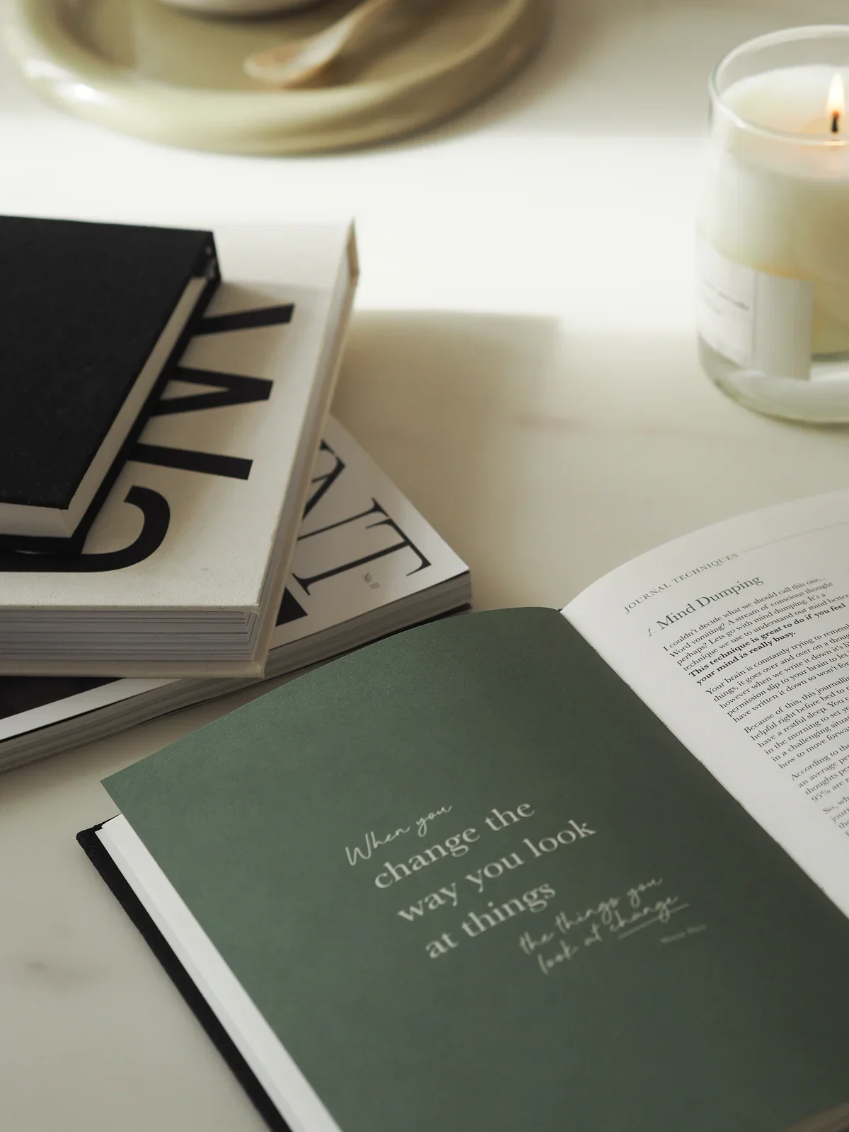 Open book with quote, closed books, candle, and a magazine or notebook on a white desk.