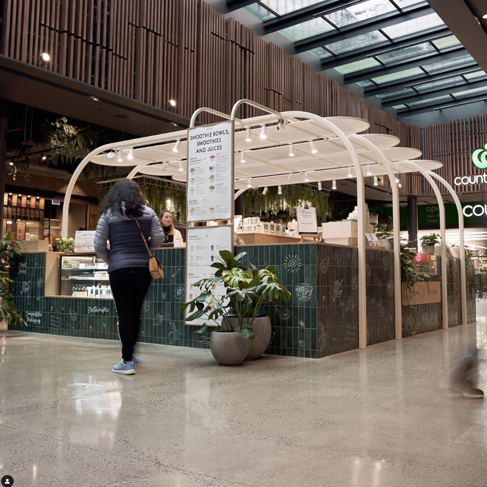 People shopping at a smoothie and juice stand inside a shopping mall with a modern design and high ceiling.