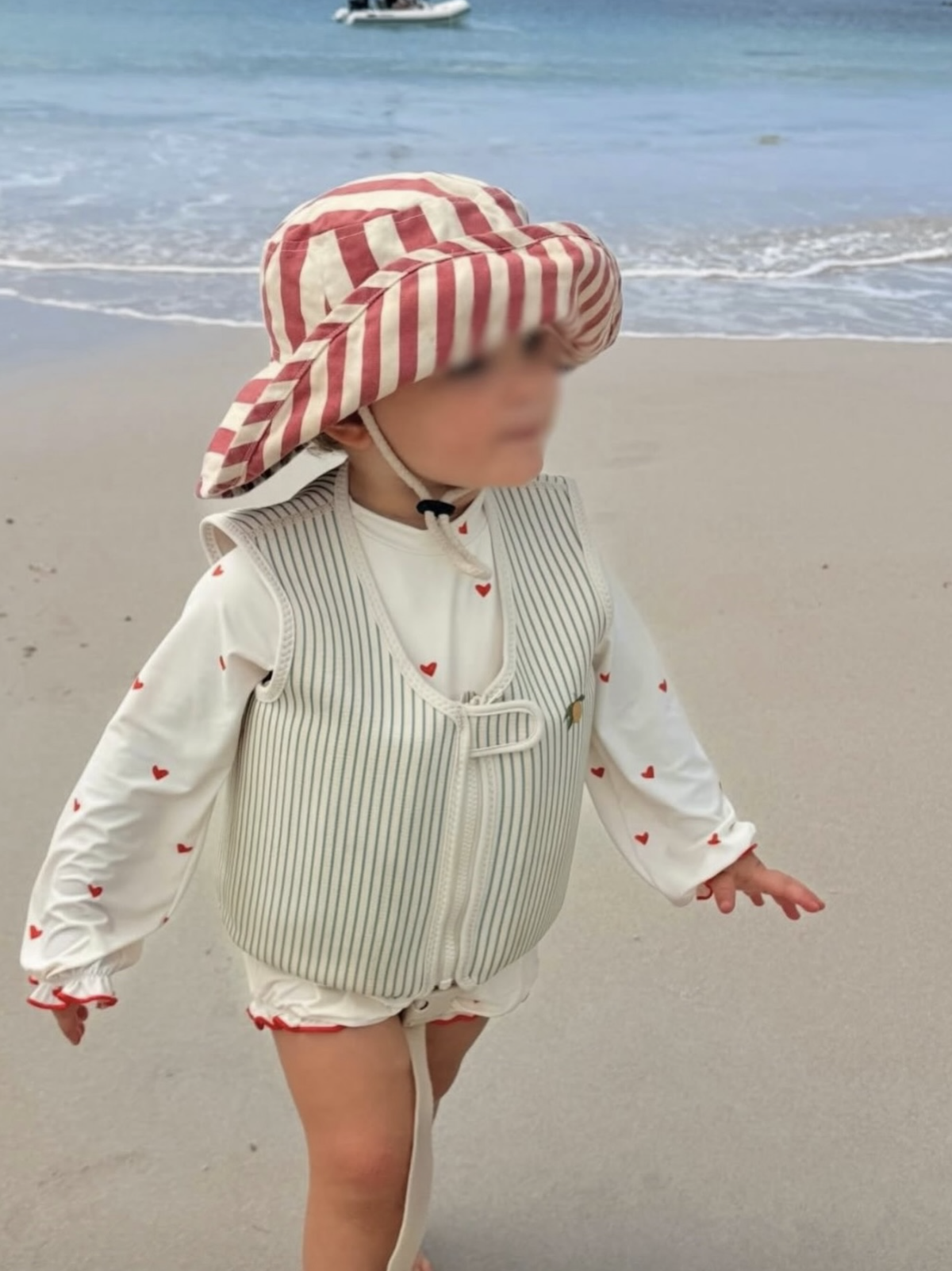 Child on beach wearing a striped wide-brim hat, white jacket with red hearts, and shorts, near the ocean with boats in background.