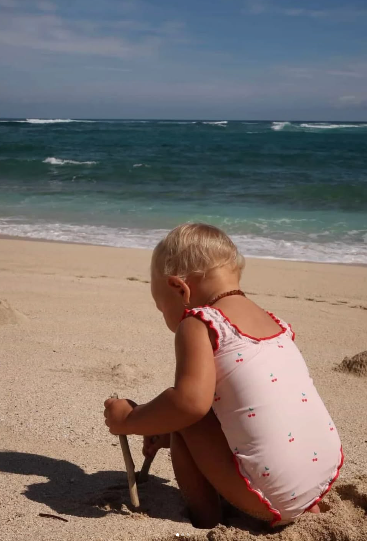 A young girl with blonde hair sitting on a sandy beach near the ocean, holding a stick, with waves crashing in the background under a partly cloudy sky.