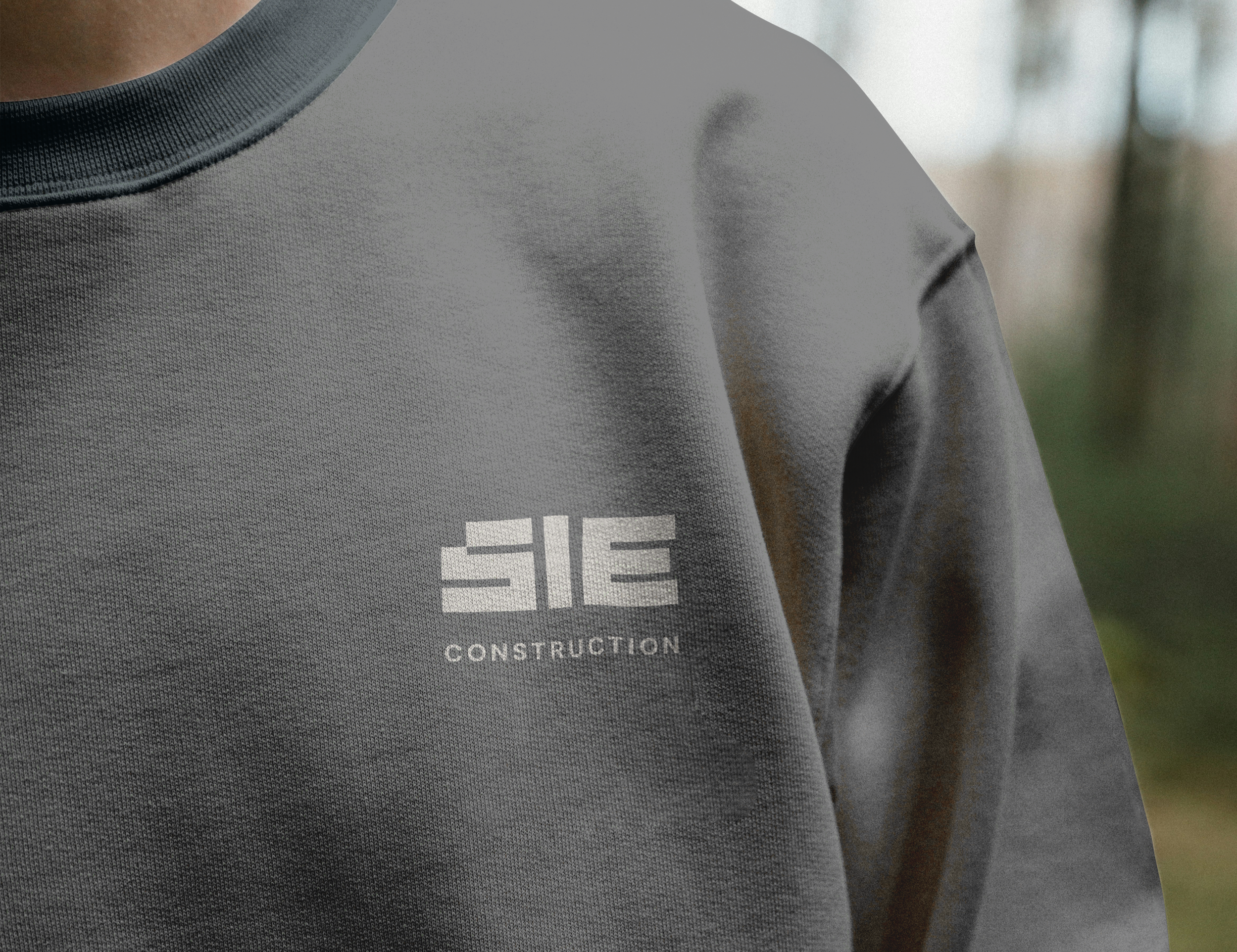 Close-up of a man wearing a gray construction T-shirt with a partially visible logo. The background is blurred with green trees.
