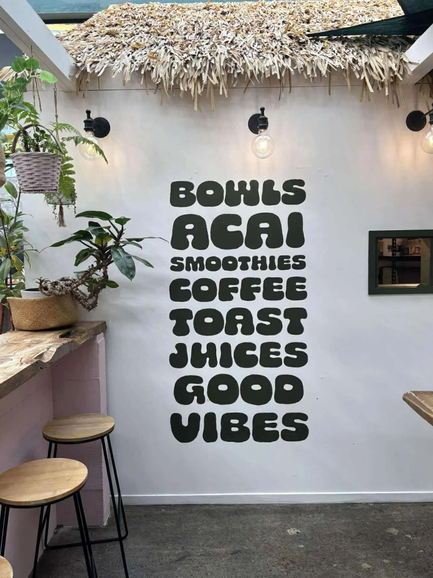 Interior of a cafe with a white wall featuring bold black text listing menu items: 'Smoothies, Coffee, Toast, Juices, Food, Vibes'. To the left, there are green plants on a wooden counter and two wooden stools with black metal legs in front of the wa