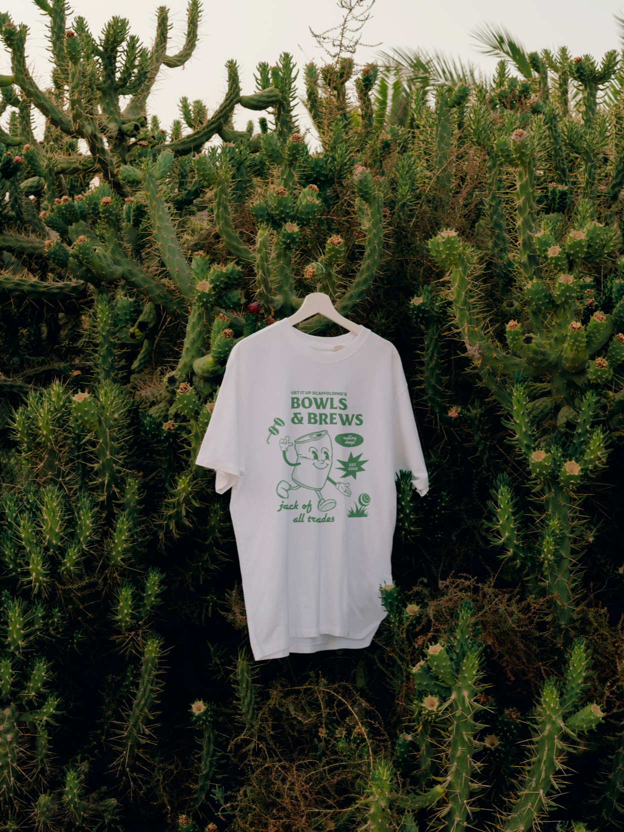 A white T-shirt with green text and illustration hanging in front of a large cactus garden. The T-shirt features a cartoon character holding a mug, with the words "Get it up scaffolding's BOWLS & BREWS Jack of all trades" printed on it.