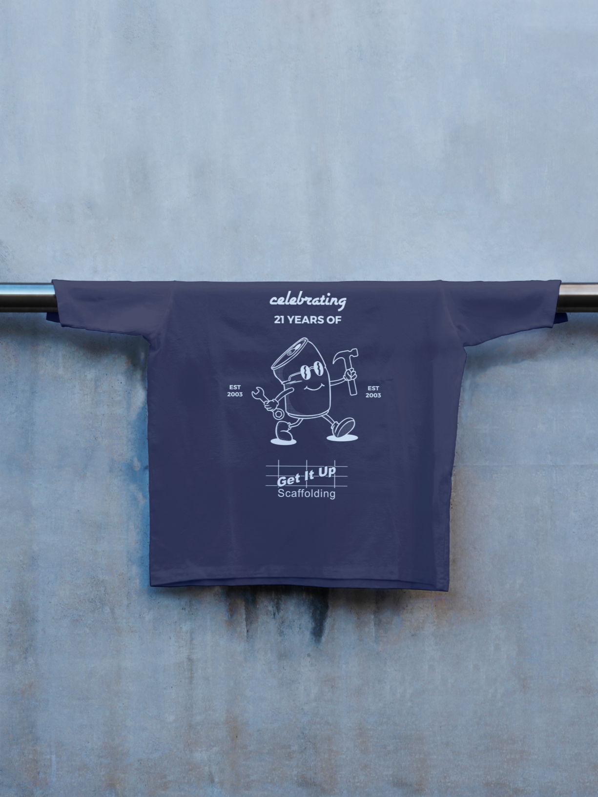 A navy blue T-shirt hanging on a metal rod against a blue wall. The T-shirt features a cartoon scaffold character holding a wrench and a hammer, with a message celebrating 21 years of scaffolding since 2003, and includes the phrase 'Get It Up.'