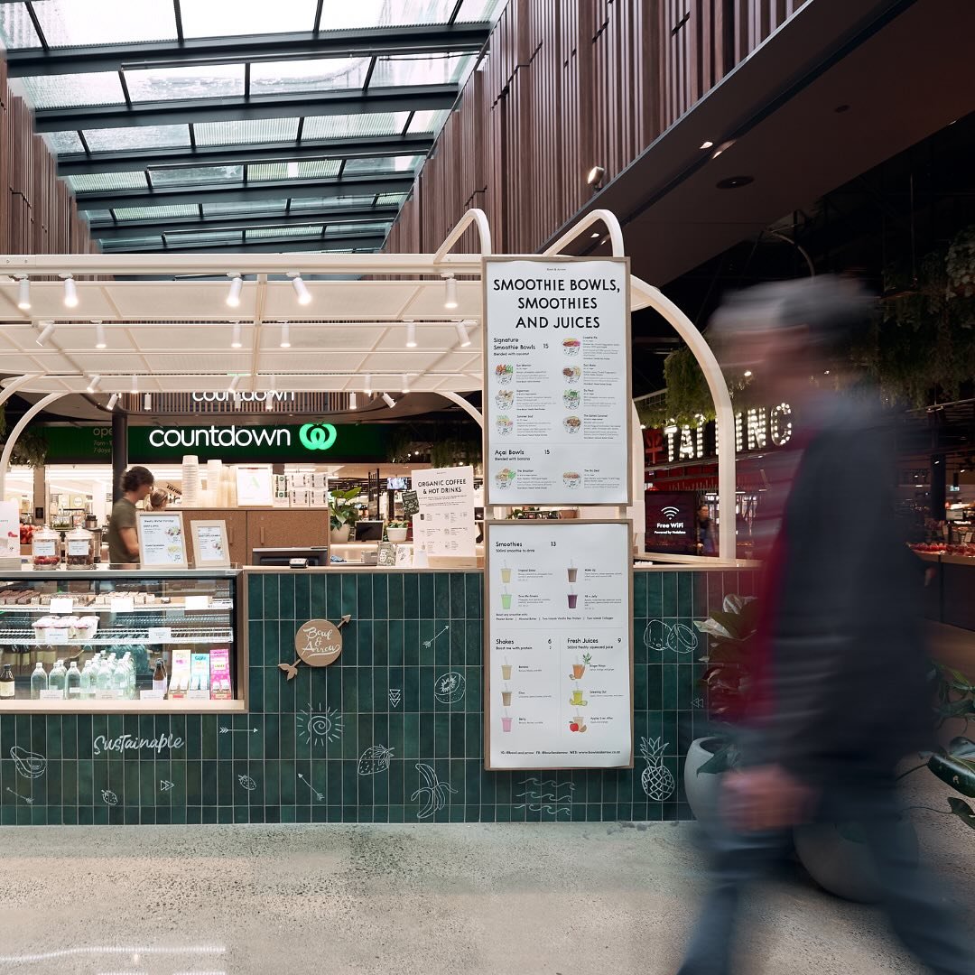 A smoothie stand in a shopping mall with a menu listing smoothies, bowls, and drinks. The stand has a green tiled front with white illustrations of fruits and the words "Sustainable" and "Bowl & Arc". In the background, there is a store named "Countd