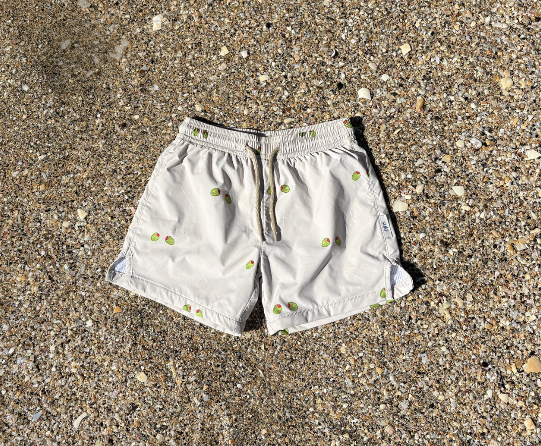 White swimming shorts with a pattern of small green and red olives laid flat on a sandy beach.