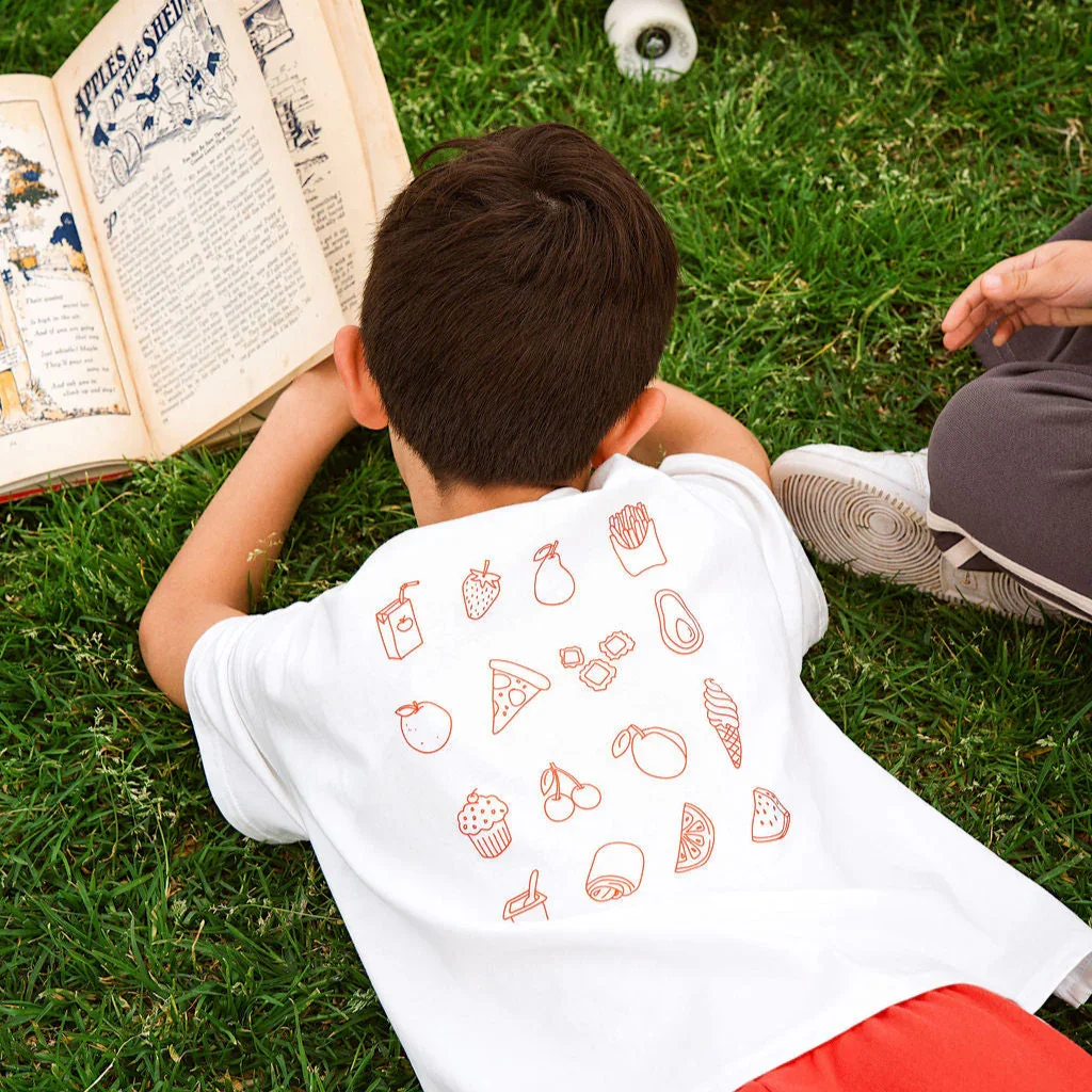 A young boy laying on the grass, reading a large open book, with a small scoop and another person's hand nearby. The boy is wearing a white T-shirt with food and drink illustrations and red shorts.