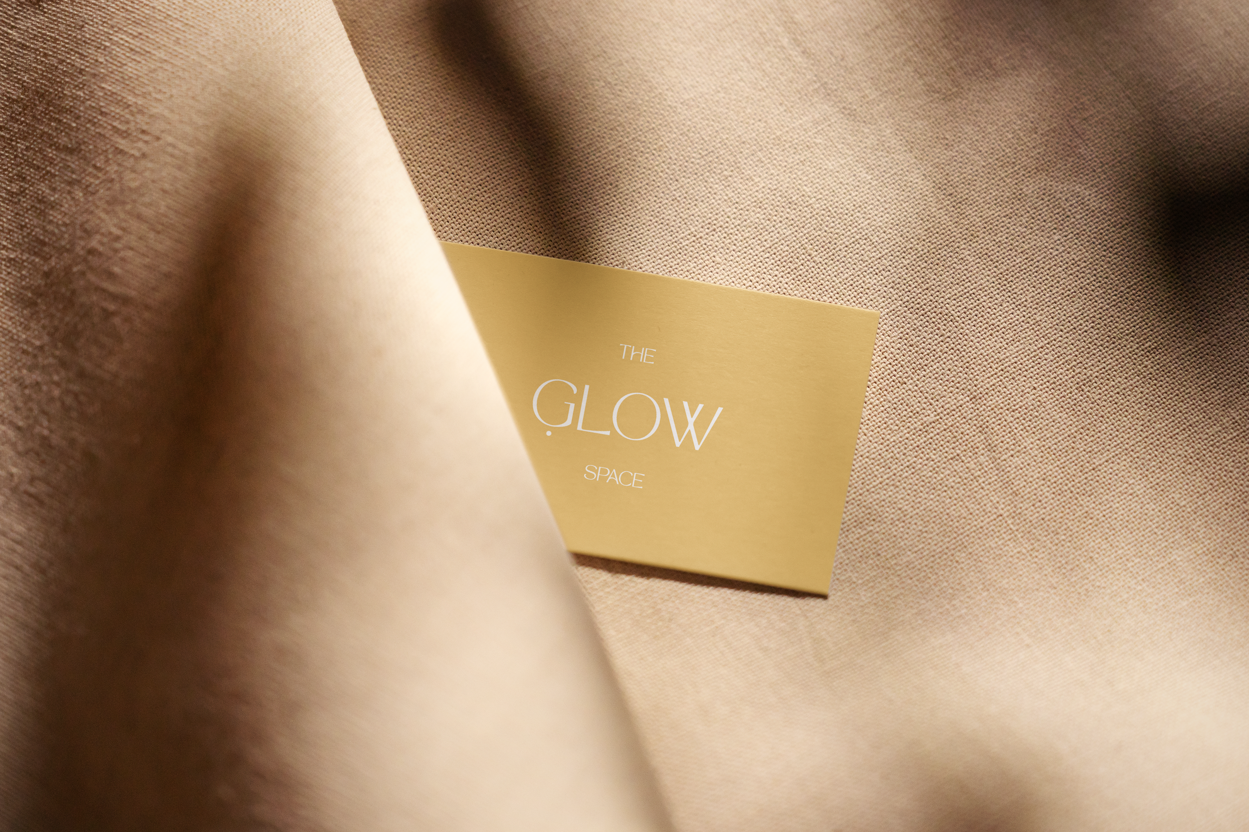 Close-up of a textured beige fabric with a pale yellow card partially visible, displaying the words 'THE GLOW SPACE' in white text.