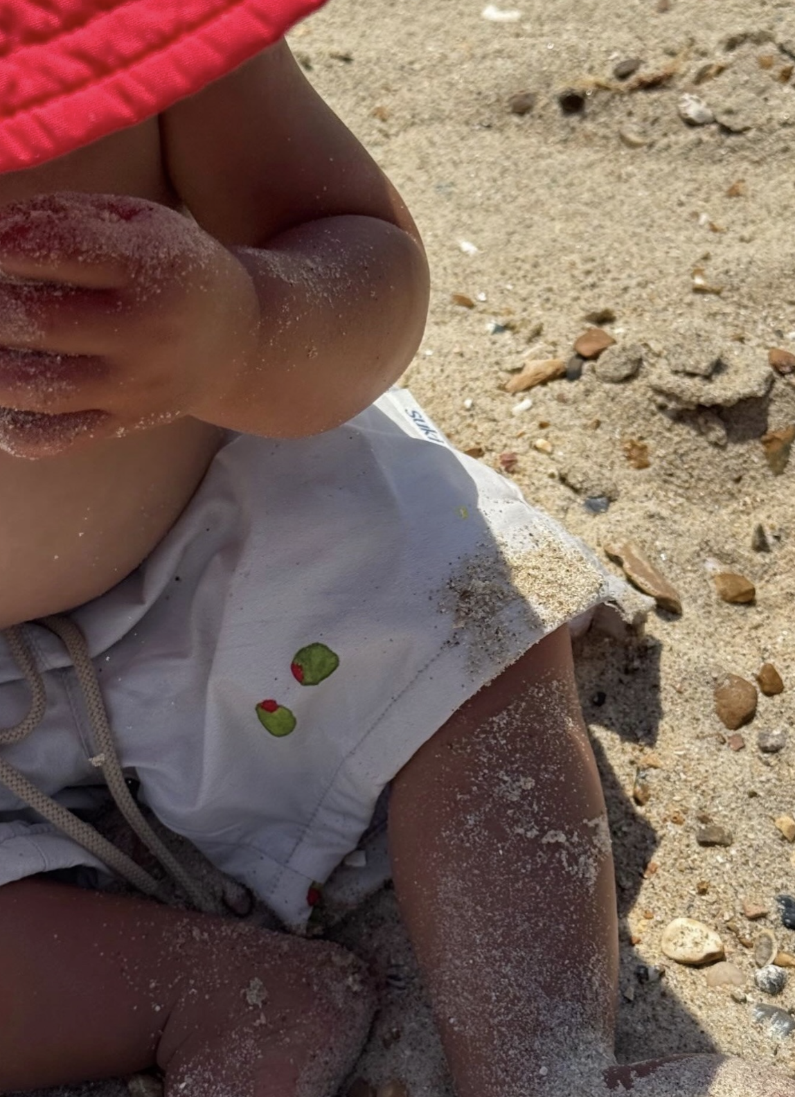 Close-up of a child's knee and part of an arm on a sandy beach, with sand on the child's skin, wearing white shorts with a small fruit pattern and a red shirt, surrounded by sand and small rocks.