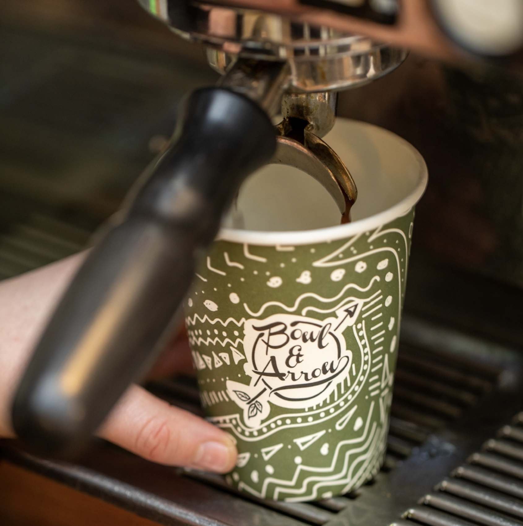 Close-up of a coffee cup with a black handle, under an espresso machine spout dispensing coffee into the cup. The cup has a green and white decorative design with the text 'Bowl & Arrow' on it.