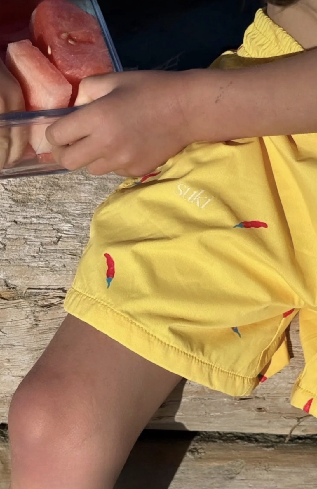 Child wearing yellow shorts with small colorful parrot print, sitting on a wooden surface, holding a container of watermelon slices.