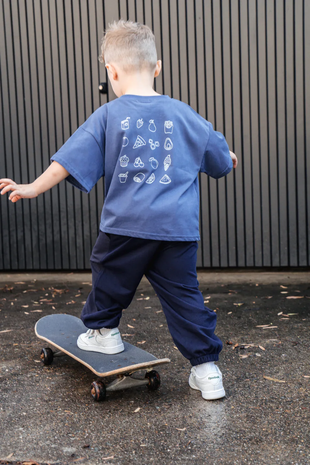 Child riding skateboard outdoors on pavement facing away from the camera, wearing a blue T-shirt with white food icon graphics on the back, dark pants, and white sneakers, with a black vertical panel background.