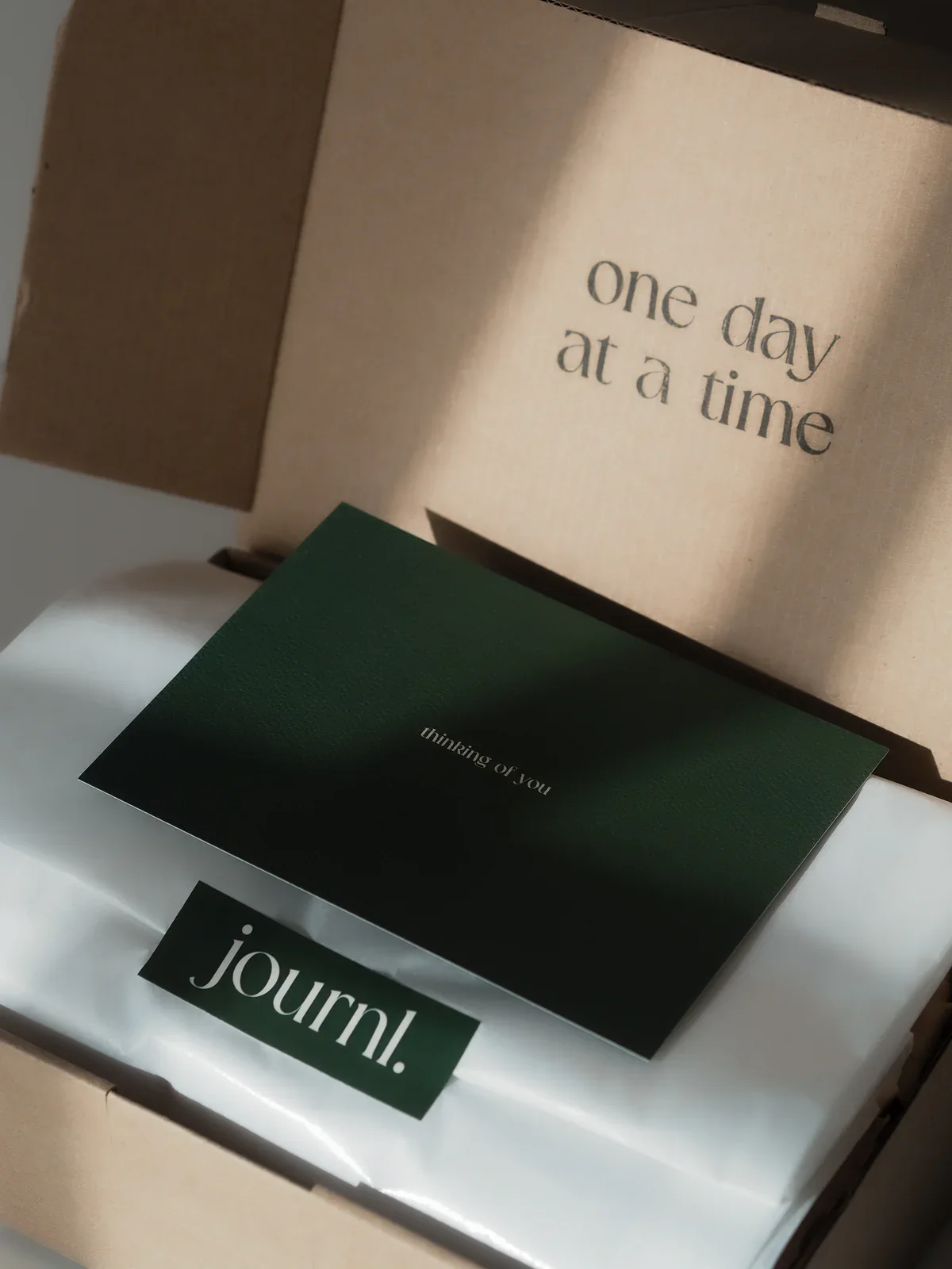A motivational message in a cardboard box that says 'one day at a time.' Below it, there is a small green card with the words 'thinking of you,' and a larger white card with the word 'journal.'