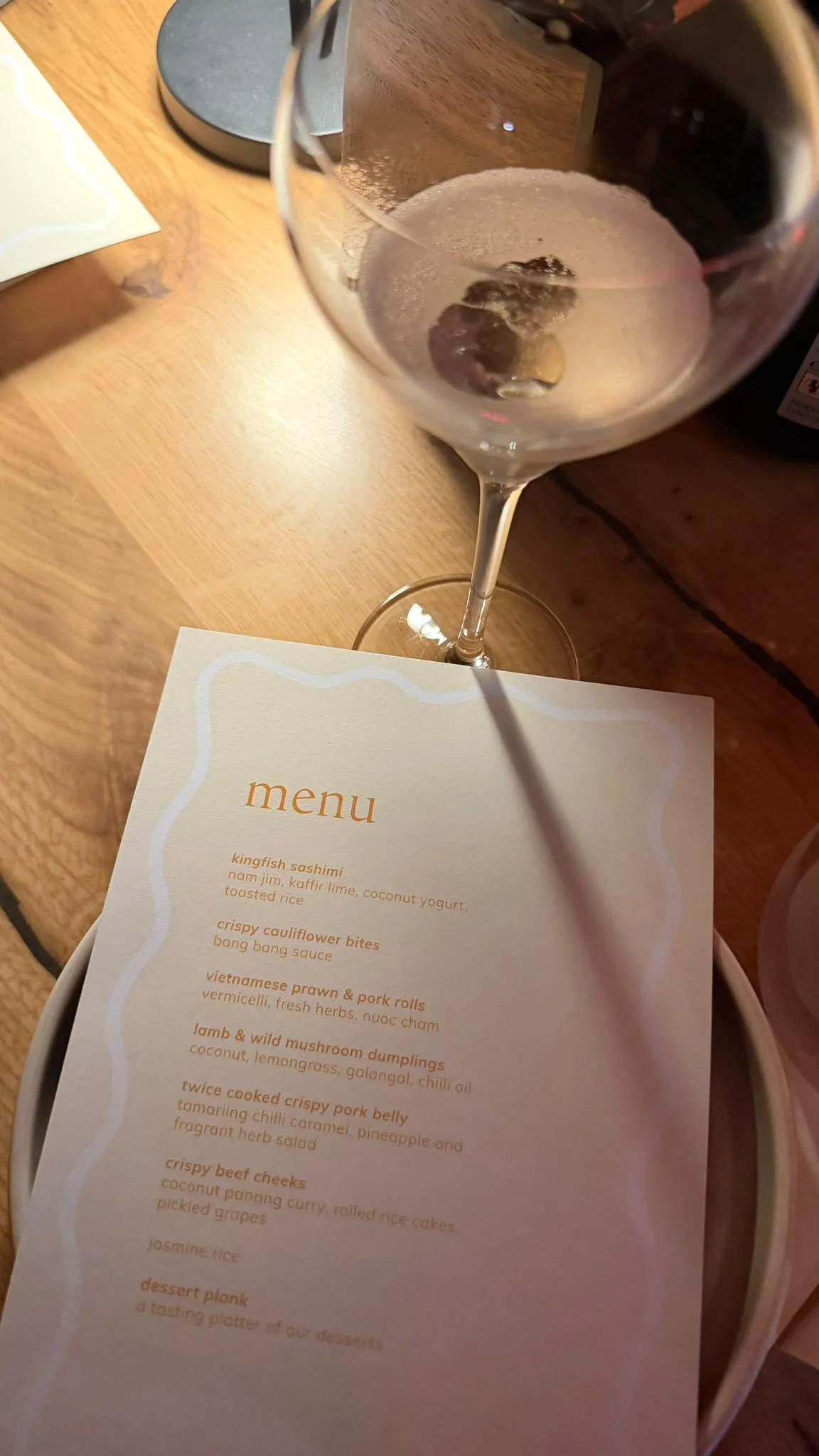 A glass of red wine on a wooden table with a menu listing various dishes.