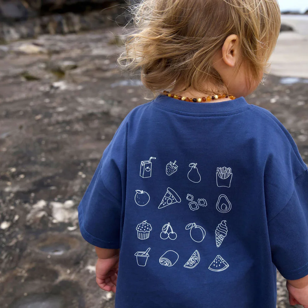 Back of a young child wearing a blue T-shirt with white line drawings of various food and drink items, including juice box, strawberries, pear, fries, apple, pizza, sushi, avocado, cupcake, cherries, peach, ice cream cone, drink cup, croissant, lemon